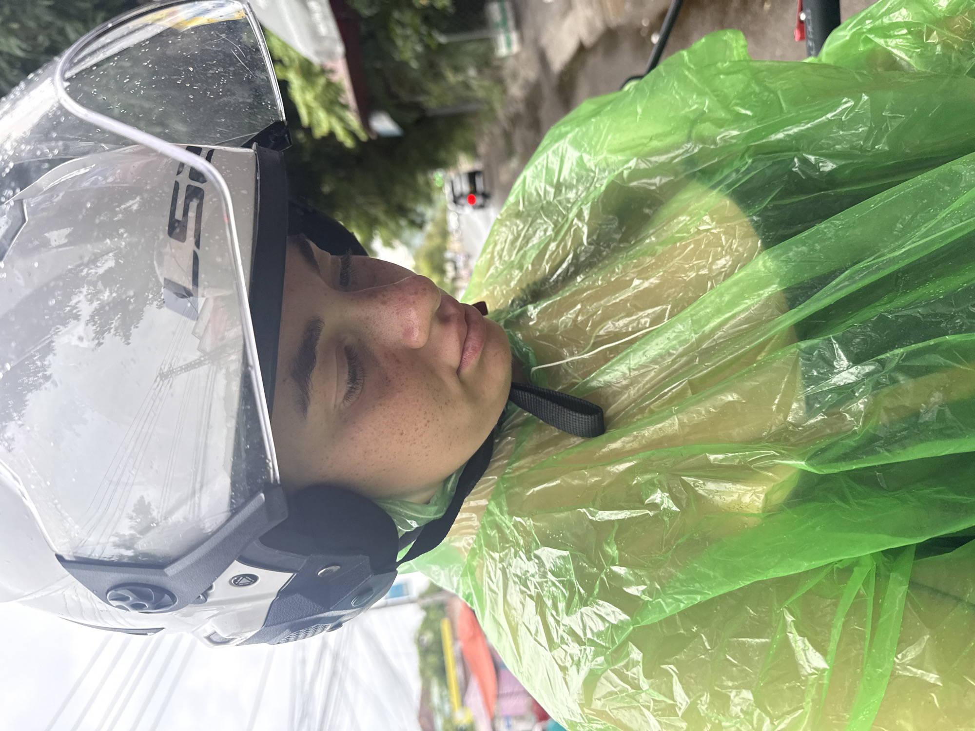 In Nha Trang, Vietnam, a person wearing a white helmet and a bright green poncho is seen looking down. The helmet has a clear visor, which is speckled with rain droplets. The poncho is made of a thin, crinkled plastic material, suggesting it is a disposable rain cover. The person's face, visible through the visor and above the poncho, shows a scattering of freckles. The background indicates an outdoor setting, likely a street, with trees, buildings, and other vehicles visible in the distance, further suggesting a rainy day in an urban environment.