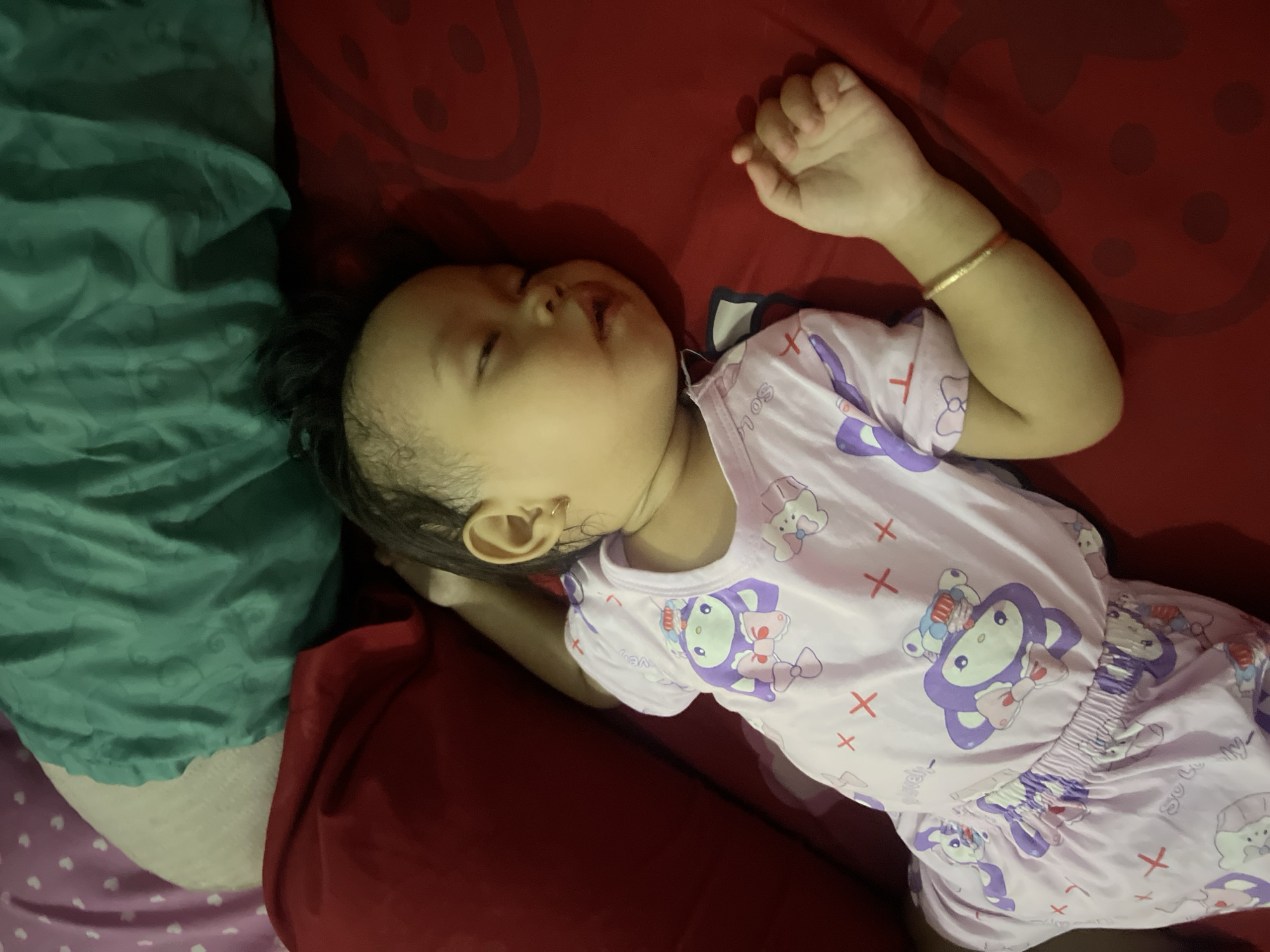 A young child, appearing to be asleep, lies on a red surface that resembles a bed. The child is wearing light purple pajamas adorned with cartoon characters, specifically what appear to be monkeys wearing bows, and other playful motifs like crosses. The child's right arm is extended outwards, and a thin gold bracelet is visible on their wrist. To the left of the child, a portion of a red pillow or blanket is visible, and behind the child, there are green and white fabrics, possibly more bedding or pillows. The overall scene suggests a domestic setting, likely a bedroom, within Kecamatan Pati, Indonesia.