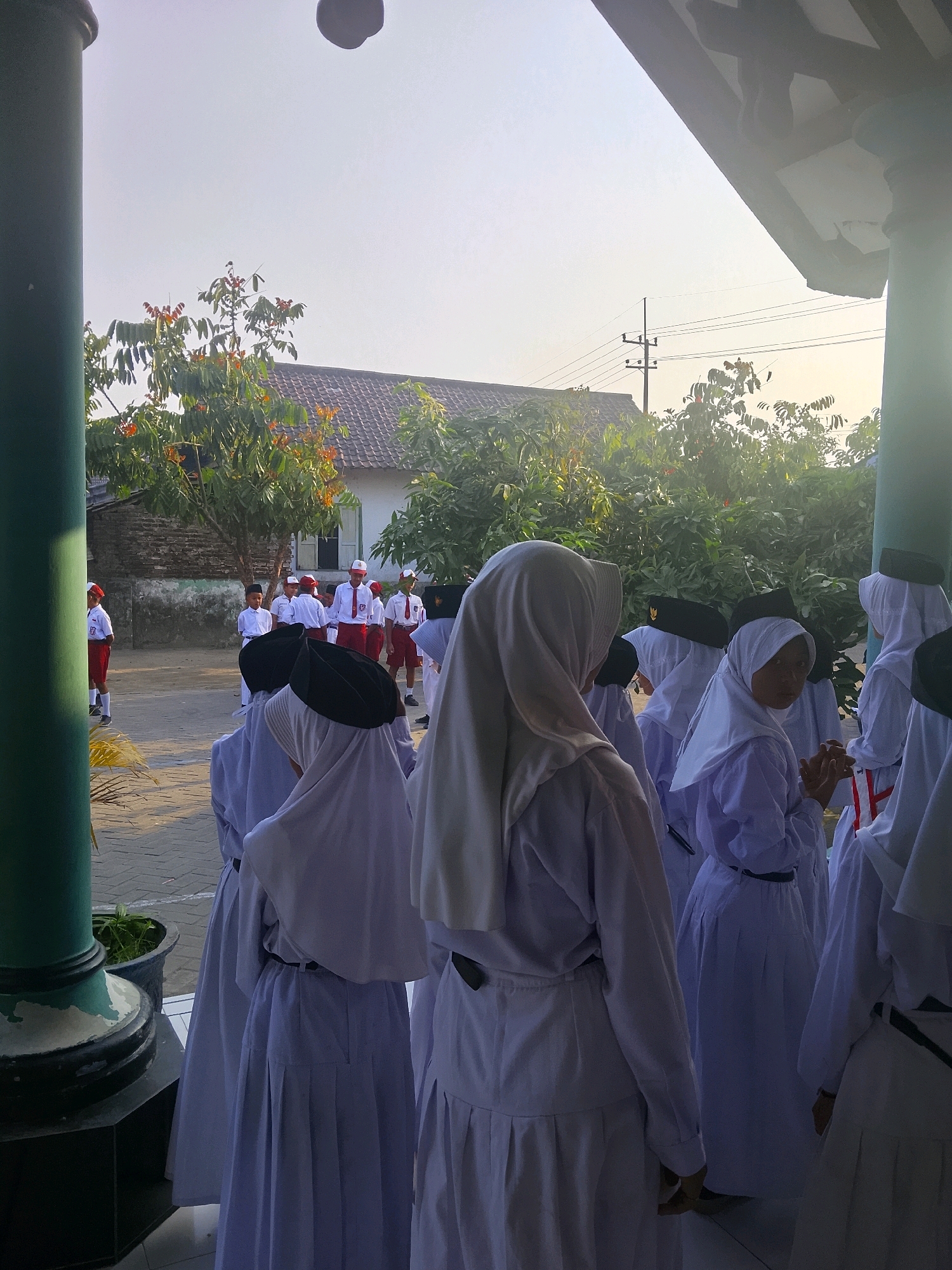 A group of students in uniform are gathered outdoors, likely at a school in Kepuh, Indonesia. The students in the foreground are wearing white hijabs and long white skirts, with some also wearing black caps. Behind them, a group of students in white shirts, red shorts or skirts, and red caps are standing. The background shows a building with a tiled roof and trees. The scene appears to be illuminated by natural light, suggesting it is daytime. The overall atmosphere is one of a school gathering or assembly.