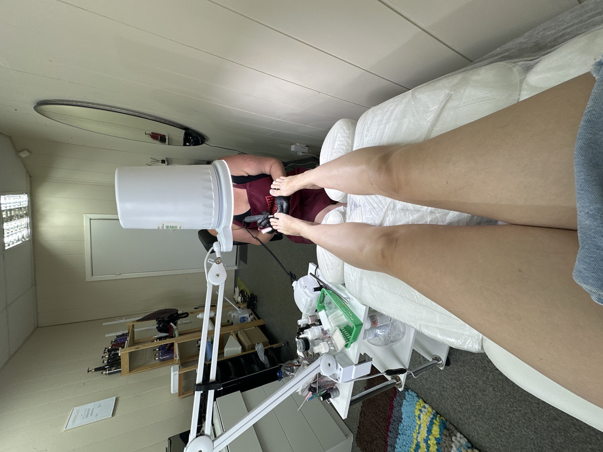 The image depicts a person receiving a pedicure in a salon in Novoaltaysk, Russia. The client's legs and feet are prominently displayed, positioned on a white treatment bed. A person, presumably the pedicurist, is bent over, wearing an apron and black gloves, and is working on the client's feet with tools, including what appears to be a nail drill. A large white bucket-like object is placed over the pedicurist's head, possibly for privacy or to shield them from some element of the treatment. The environment appears to be a small salon with shelving units stocked with bottles, a mirror on the wall, and fluorescent lighting. A white wheeled cart holding various manicure and pedicure supplies is situated next to the treatment bed, equipped with a lamp and numerous bottles and containers. The flooring is carpeted with a colorful rug visible in the lower left corner.