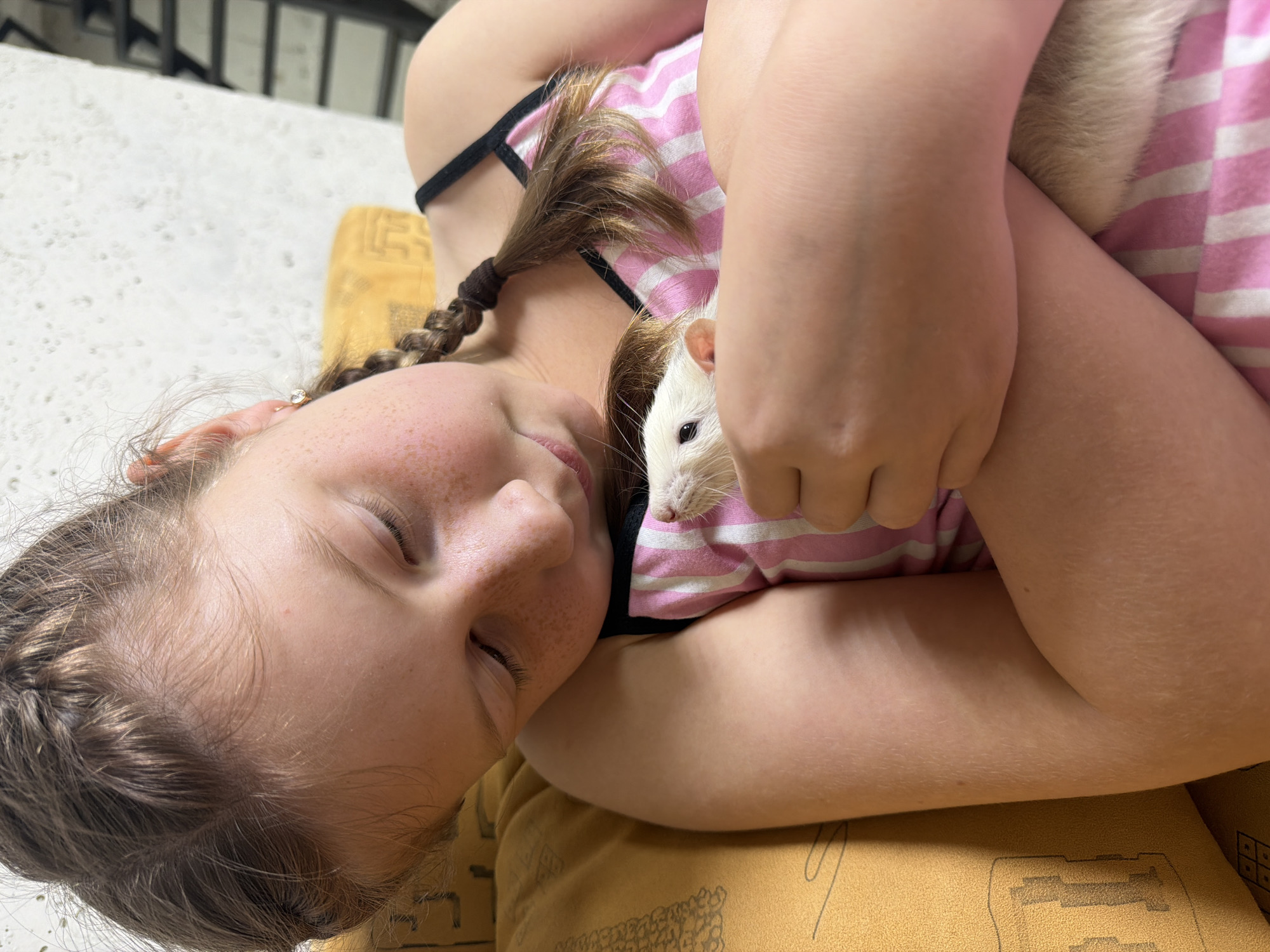 A young fair-skinned girl with freckles, eyes closed, and braided hair lies on a yellow patterned surface. She wears a pink and white striped tank top with black straps and is cradling a white rat against her chest. The rat's head is visible, with a pink nose and black eyes. The background is blurred, showing a pale surface with vertical lines. There is no evidence to suggest the location is Markovo, Russia.