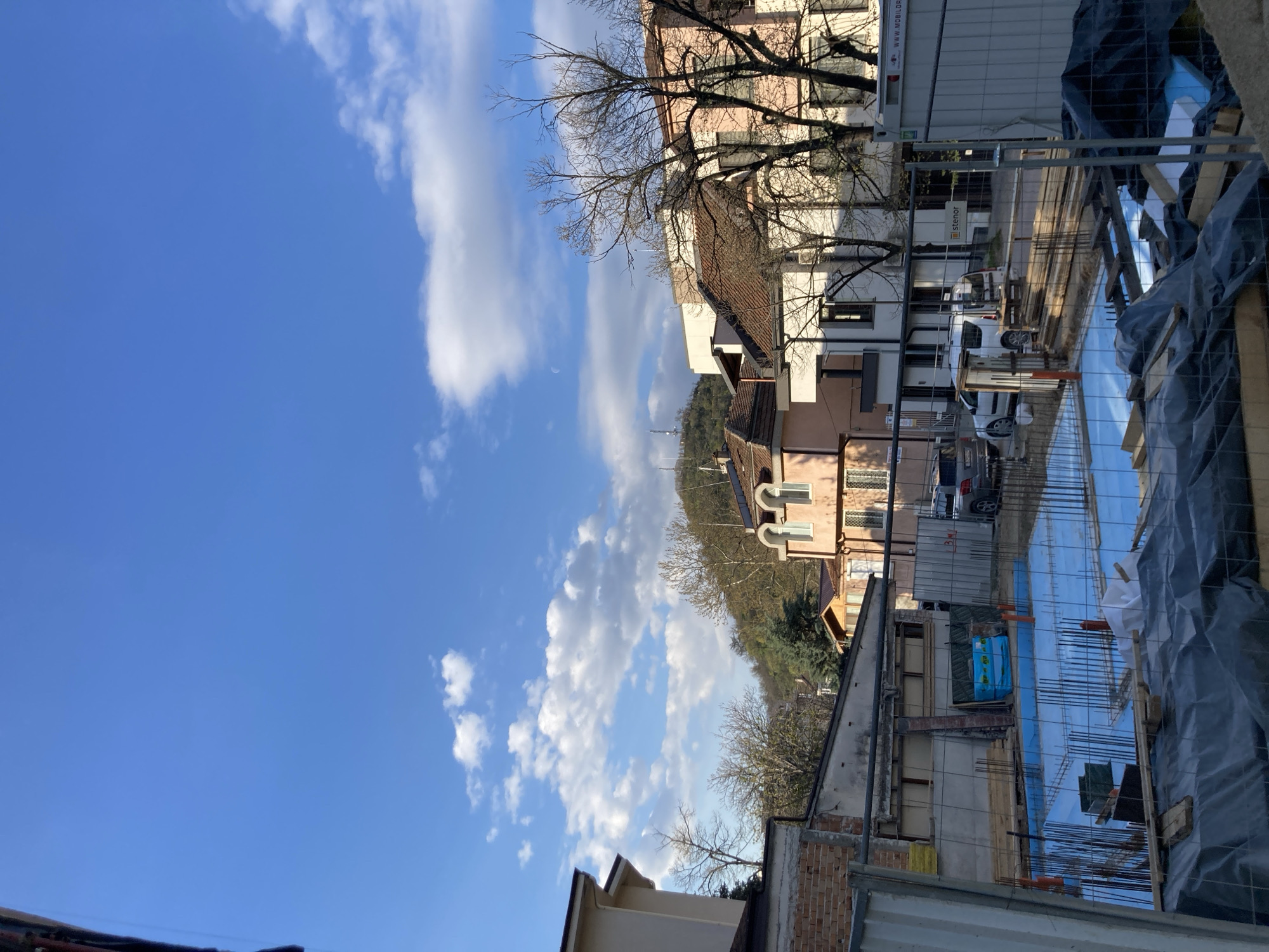 The image depicts a street scene in an urban area. Buildings line the street, including residential homes and apartment complexes. Several vehicles are parked along the street. A construction site is visible, with rebar and protective sheeting covering the ground. The sky is predominantly blue with scattered clouds. Bare trees are interspersed among the buildings.