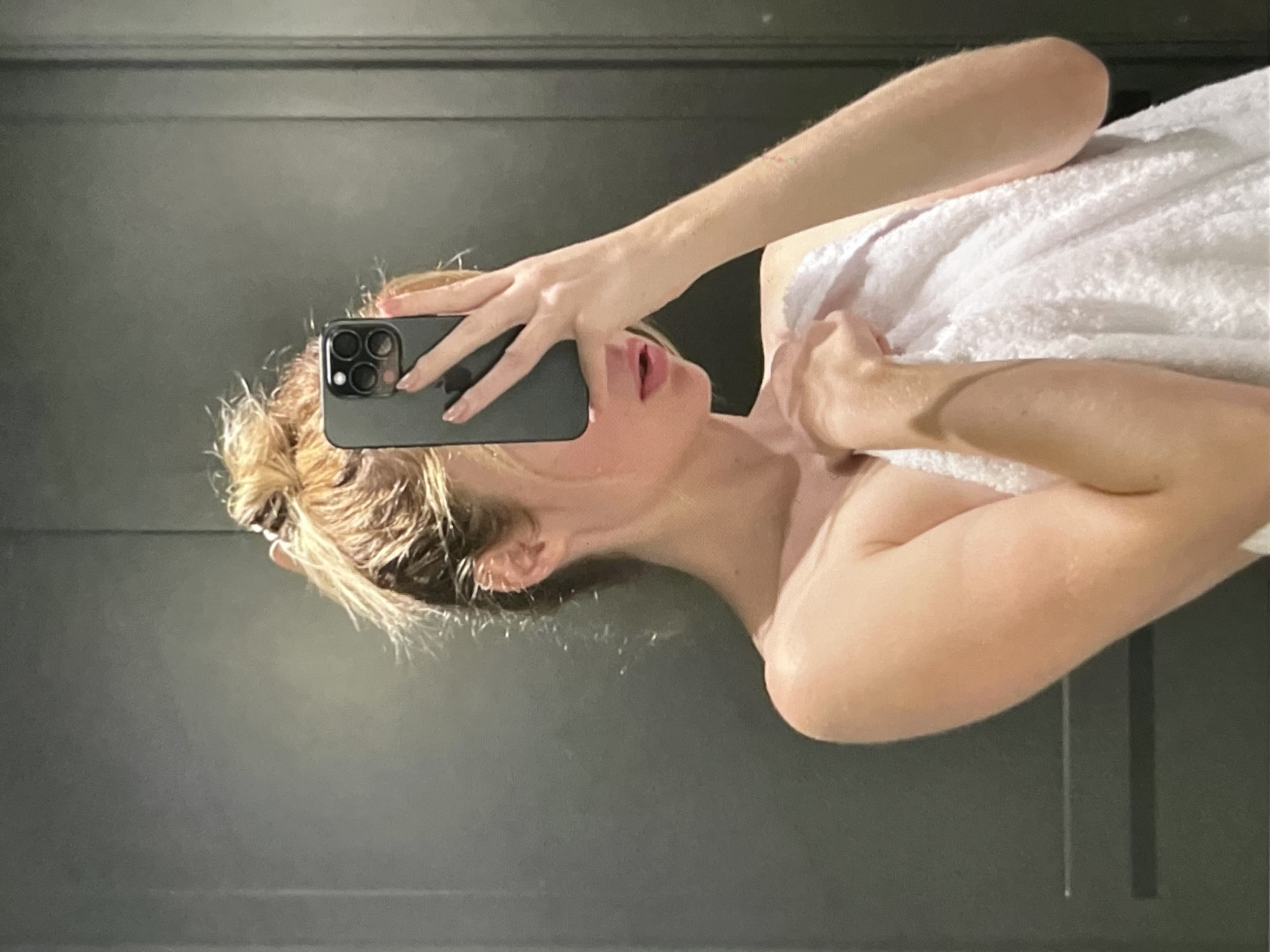A person, appearing to be female with blonde hair pulled up in a messy bun, takes a selfie in a mirror. She is wearing a white towel wrapped around her body and is holding a smartphone in her right hand, obscuring her face. The background appears to be a bathroom with dark gray walls or doors. The lighting is dim, casting soft shadows. The image is a close-up shot, focusing on the upper body and face of the person. The location is Unknown, Unknown.