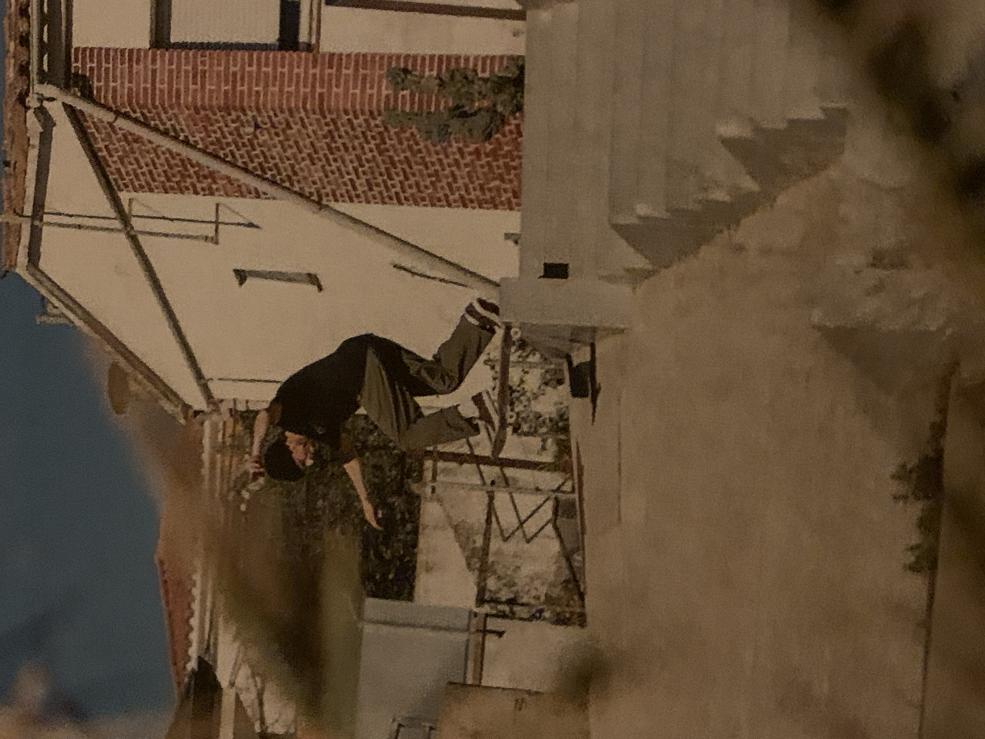A person on a skateboard is captured in motion mid-air, appearing to jump off a low wall. The individual is wearing dark pants and a dark shirt. The setting is an urban environment, with a building featuring a brick roof and white walls in the background. Below the skater, there are chairs and a fence. The backdrop includes another building and what appears to be sky. The image provides no indication about the specific location in Barcelona.