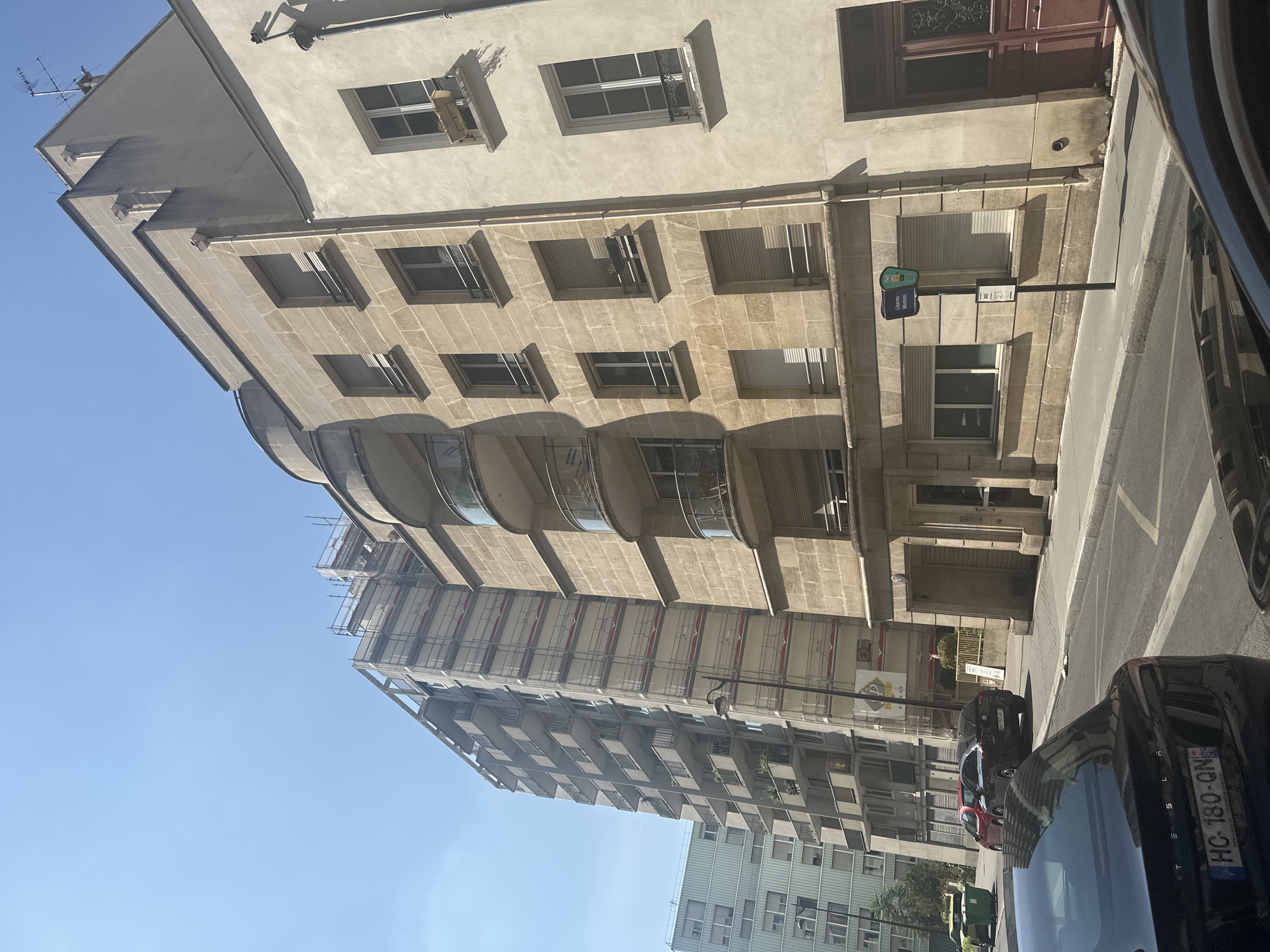 The image depicts a street scene in Paris, France, with a focus on several apartment buildings. On the right, a building with beige stone facade and rounded balconies with glass railings is visible. To its left, a taller apartment building is under renovation, with scaffolding covering a significant portion of its facade. Further to the left, a modern apartment building with a light green facade and numerous windows stands. In the foreground, several cars are parked on the street, including a black Tesla with a license plate that reads "HC-180-QN". A red car and a black SUV are also visible in the street. The environment appears to be urban, with a clear blue sky overhead and sunlight casting shadows on the buildings. A street sign indicating "Charles Michels" is mounted on a pole on the sidewalk.