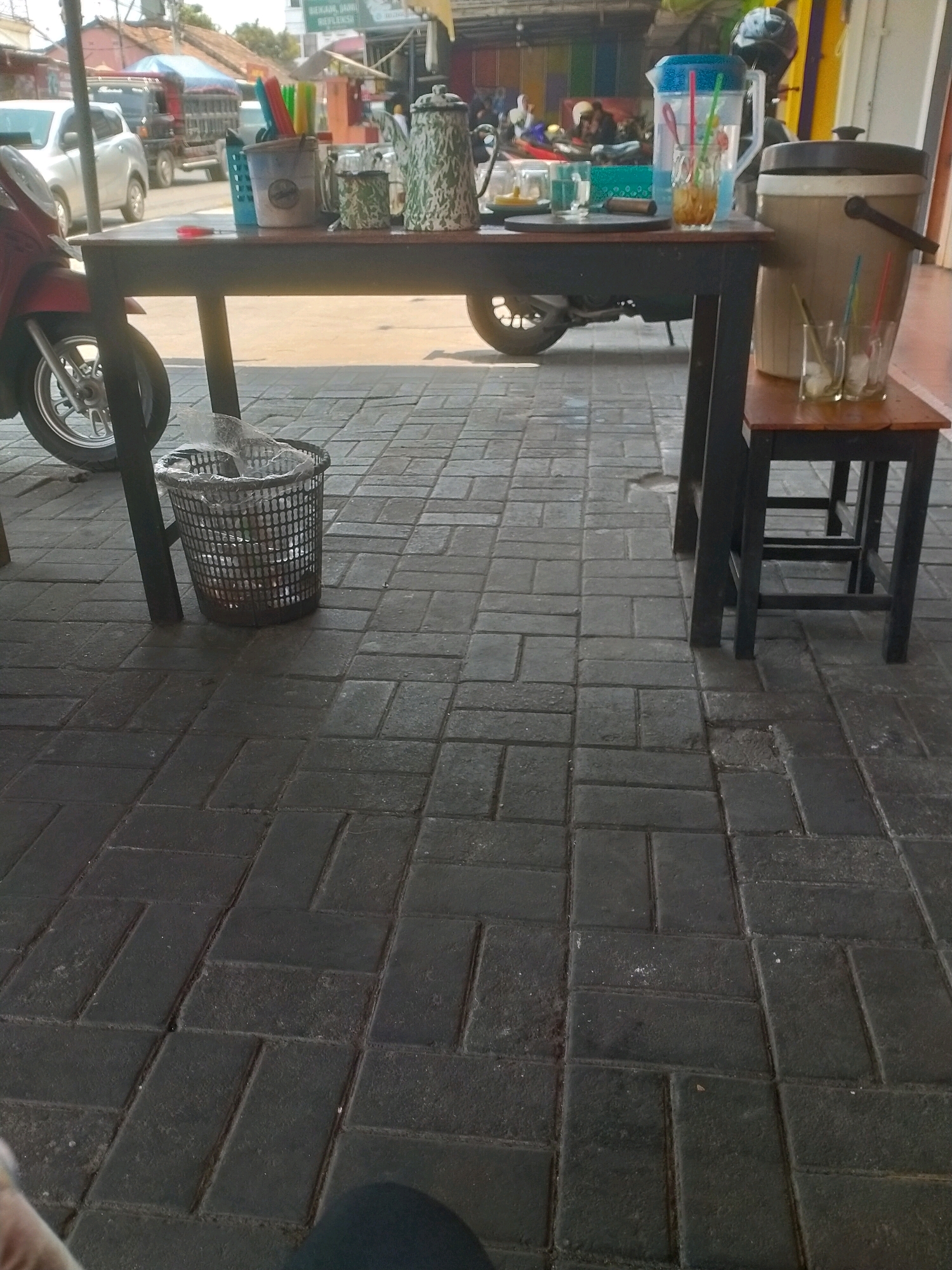This image captures a scene on a paved sidewalk in front of a shop or stall in Kasihan, Indonesia. The foreground is dominated by dark gray, rectangular paving stones arranged in a running bond pattern. To the left, the front wheel and fender of a red motorcycle are visible. A black wire trash can, filled with clear plastic bags and some debris, sits on the paving stones.
In the midground, a dark-colored wooden table with black legs is positioned on the sidewalk. On the table are several items: a blue plastic basket containing colorful straws, two glass jars, a patterned metal teapot, and a smaller patterned metal container. To the right of the table, a small wooden stool with a brown top is present. On the stool are three clear glasses, each containing ice and a straw, and a large, light-colored insulated beverage dispenser with a dark lid. A red plastic object, possibly a lid or cap, is on the ground near the table.
The background shows a street scene with vehicles, including a silver car and a truck, and structures that appear to be part of a marketplace or commercial area, including colorful awnings and building fronts. The lighting suggests it is daytime.