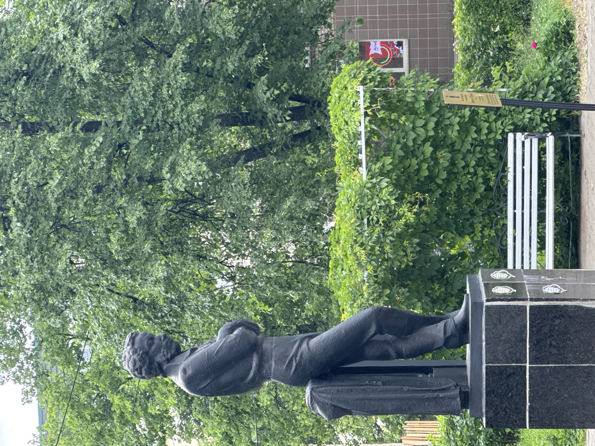 The image depicts a full-body statue of a man with his arms crossed, standing against a backdrop of lush green trees. The statue is dark in color and appears to be made of metal. It is mounted on a multi-tiered black granite pedestal adorned with what seem to be decorative metallic emblems. To the right of the statue, a white park bench with a slatted seat and back is visible, partially obscured by thick green foliage. In the background, a building with brown tiled walls can be seen, with a window displaying a neon sign. The overall environment suggests an outdoor urban setting, possibly a park or a public square, in the city of Chaikovsky, Russia. The lighting indicates it is daytime, with overcast skies contributing to a diffused illumination. The image is captured from a slightly low angle, emphasizing the height of the statue.
