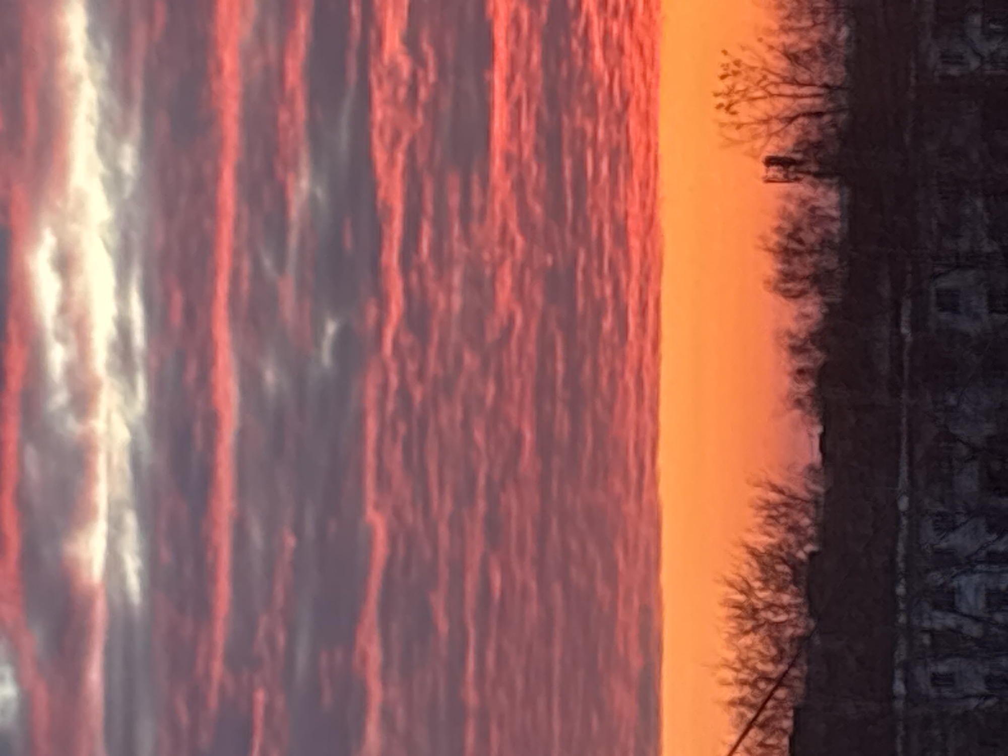 This image depicts an urban landscape in Chaikovsky, Russia, at either sunrise or sunset. The upper two-thirds of the frame are occupied by the sky, featuring multiple horizontal cloud formations. These clouds display a primary coloration of deep red and fuchsia, interspersed with bands of dark grey-violet and occasional streaks of pale yellow-white light in the upper left and right regions. Directly above the horizon, a distinct, unbroken band of intense orange light extends across the entire width of the image. The lower third of the scene consists of a dark silhouette representing the city's terrain. This silhouette includes the upper portions of several low-rise buildings, indistinct in detail, and numerous bare deciduous trees, their branching structures visible against the bright horizon. A diagonal dark line, consistent with an overhead utility wire or cable, crosses the upper-left area of the silhouette. To the right of the image's center, a vertical, dark structure resembling a telecommunications mast or antenna array is silhouetted against the orange sky, rising above the tree line. No individuals, animals, or active interactions are present within the frame.