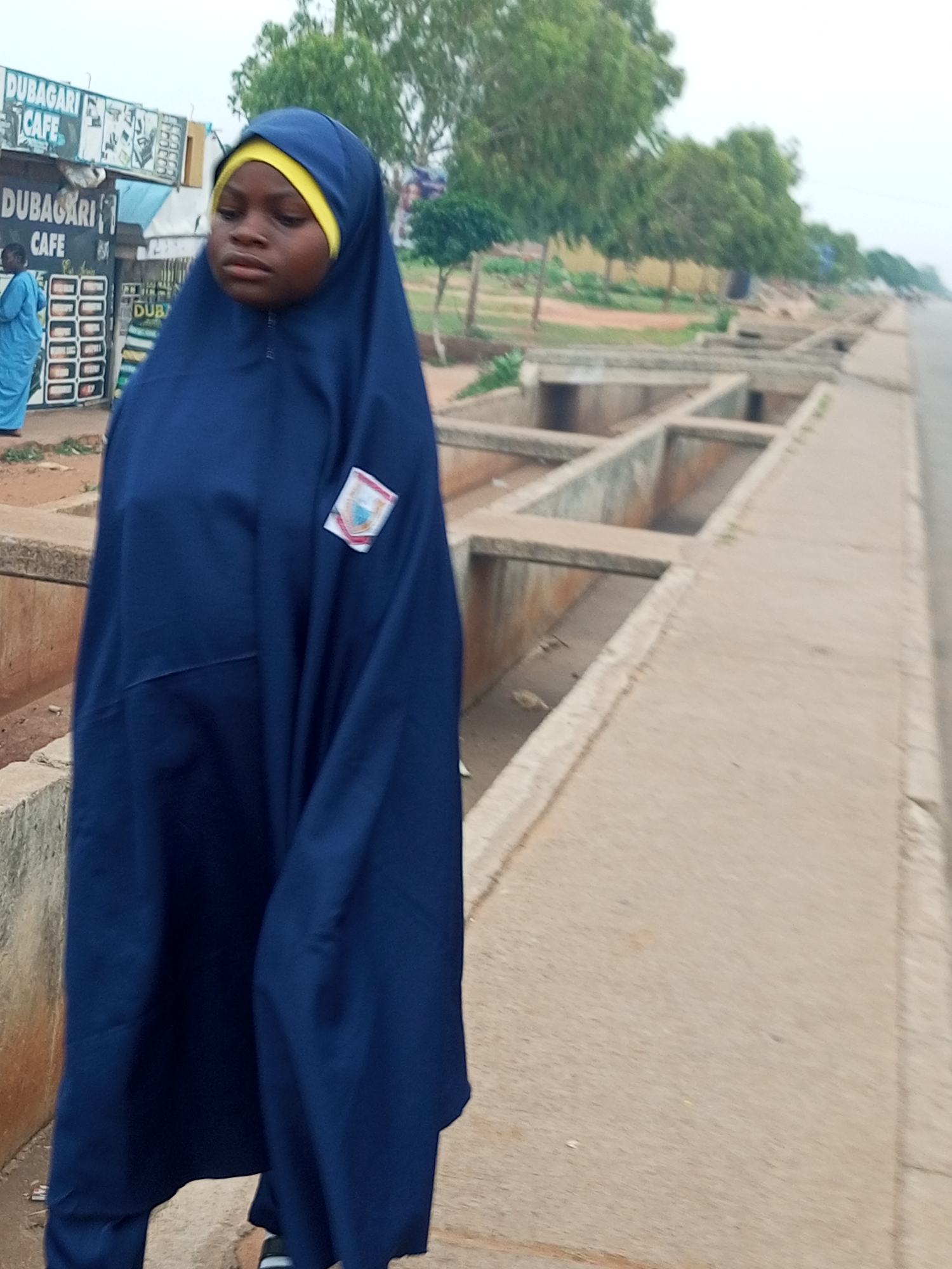 A young girl wearing a dark blue hijab and yellow head covering stands on a paved walkway in Wuro Dole, Nigeria. She is looking down and to her left. A building with signs that read "DUBAGARI CAFE" is visible behind her. The walkway is next to a concrete drainage system, and a road is to the right of the walkway. Trees line the other side of the road. The overall atmosphere is overcast.