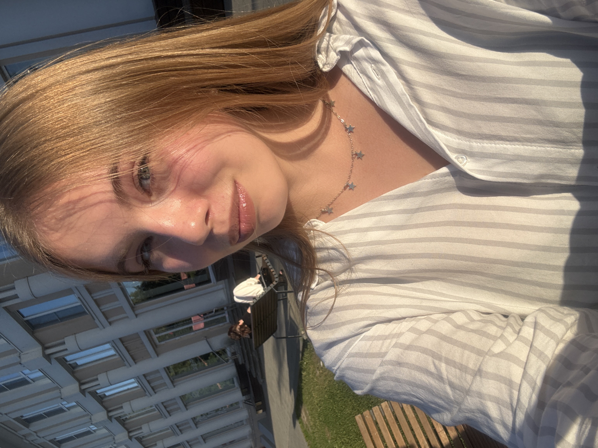This is a close-up, slightly angled selfie of a young Caucasian woman with blonde hair. She is wearing a white and light grey striped button-up shirt and a delicate silver necklace with small star charms. Her face is tilted, with one eye visible, showing blue-green irises. Her lips are slightly parted and have a glossy sheen, suggesting lip gloss or balm. Sunlight casts a warm glow on her skin, creating highlights and soft shadows, indicating it is daytime.
The background is out of focus but appears to be an outdoor setting. In the upper left, there is a glimpse of a modern architectural structure with a glass roof and visible beams, suggesting an atrium or a covered walkway. Further in the background, there are hints of greenery and perhaps other people, but they are indistinct. The overall impression is of a casual, sunny day. The image does not contain any visible text. The subject's expression is serene and relaxed.