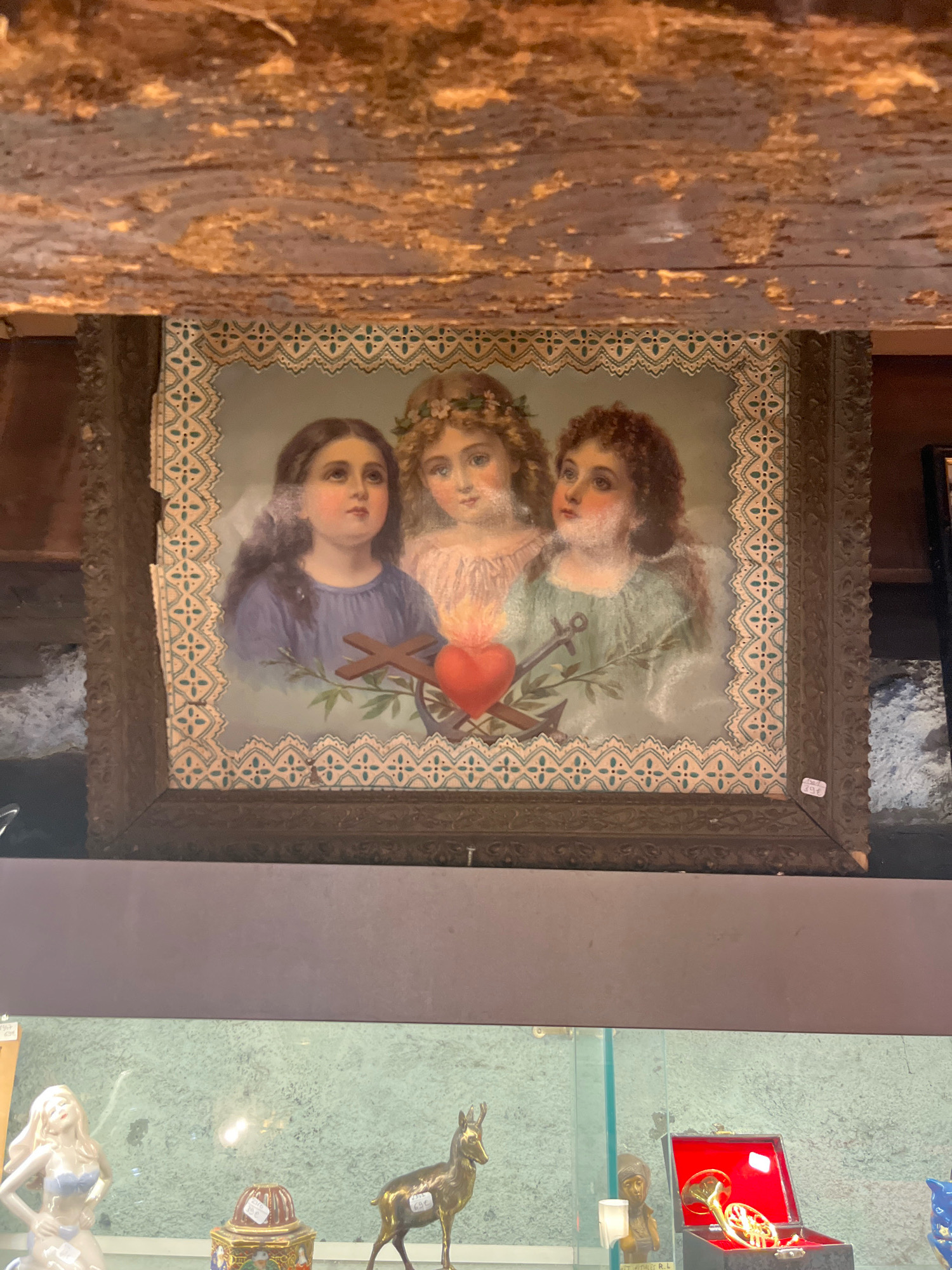 In Yvoire, France, an antique painting is displayed under a rough, textured ceiling. The painting, framed in dark, ornate wood, depicts three young figures with angelic features, surrounded by a decorative border. In the center, a vibrant red heart is pierced by two crosses and flanked by anchors, suggesting religious or symbolic meaning. Below the painting, a glass display case exhibits various decorative items, including a porcelain figurine of a scantily clad woman, a decorative box with intricate designs, a metal sculpture of a deer, a small statue of a person, and a red jewelry box containing a French horn. The overall scene suggests an antique shop or a curated collection of historical artifacts.