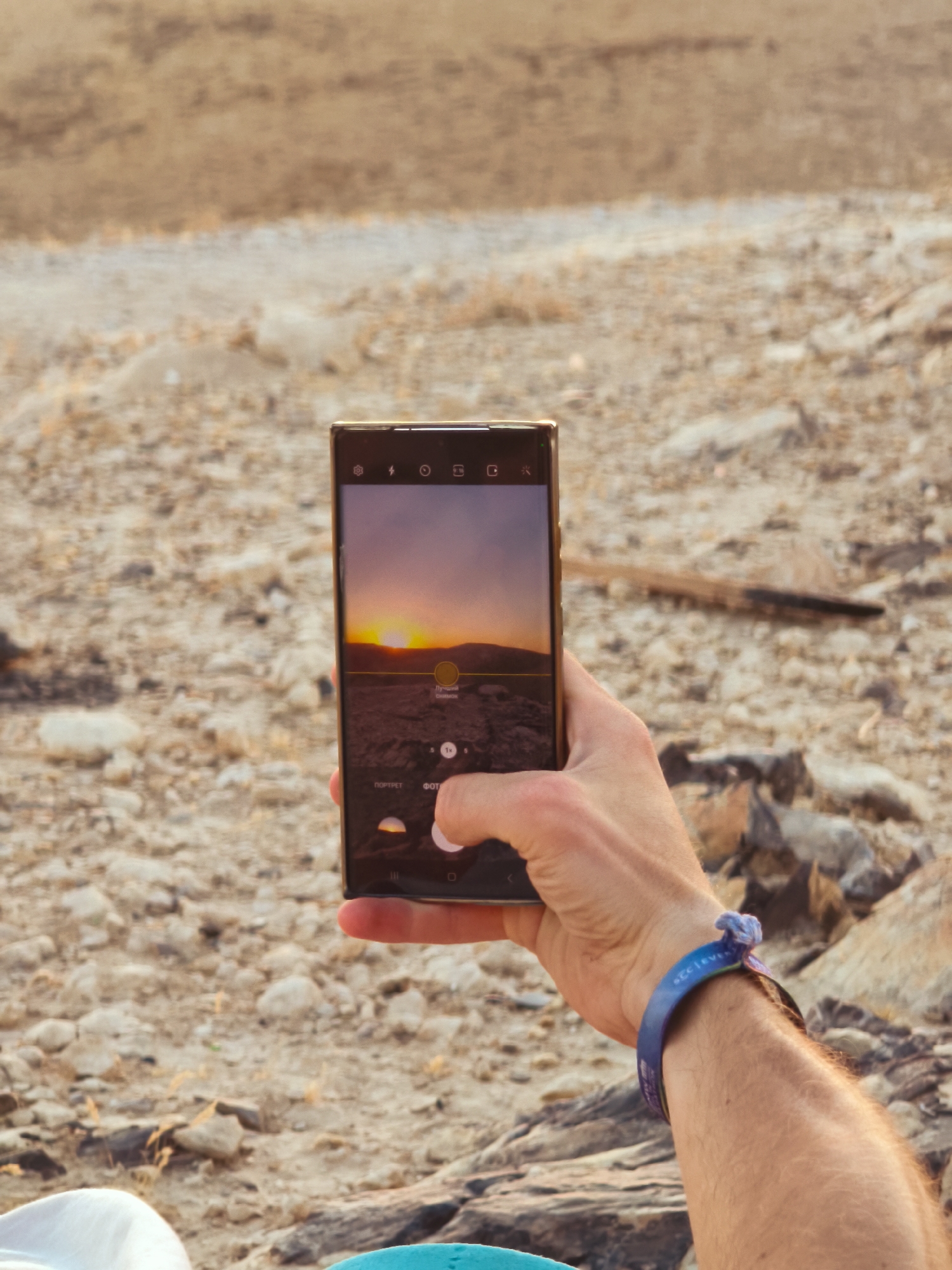 A person's hand holds a smartphone, capturing a sunset over a rocky, arid landscape. The phone screen displays a scenic view with the sun low on the horizon, casting an orange glow. The background consists of dry earth and scattered rocks, suggesting an outdoor, possibly desert or wilderness setting. The person is wearing a blue wristband with text. Part of another person wearing a white shirt is visible in the lower left corner. The overall lighting suggests late afternoon or early evening.