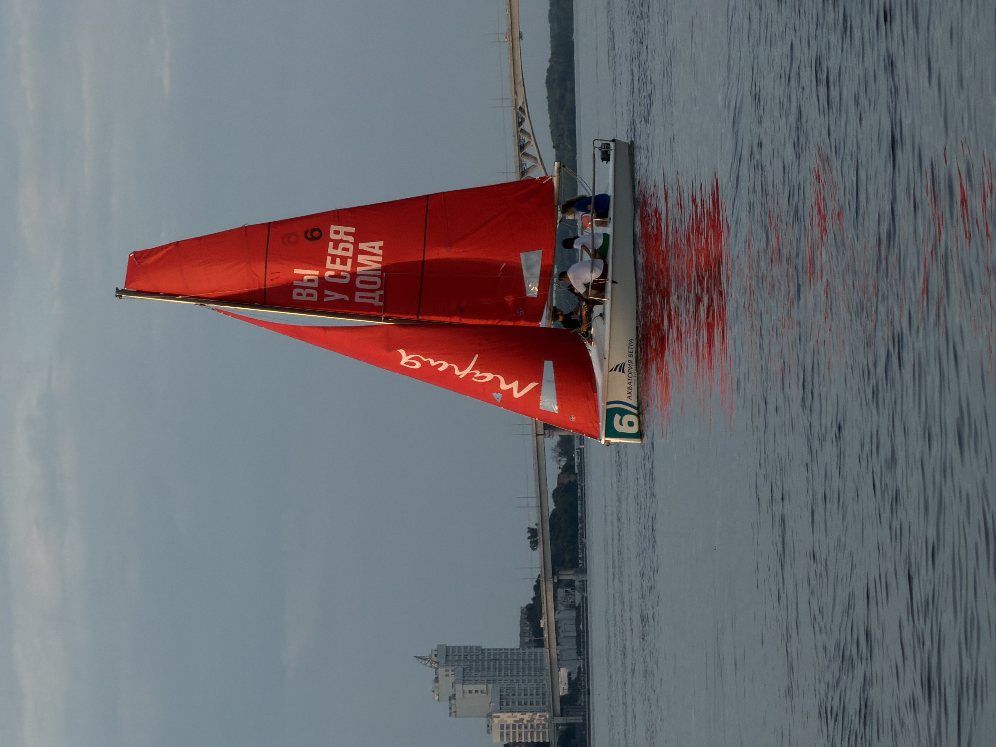A white sailboat with a red sail is on the Volga River in Saratov, Russia. The sail has white Cyrillic lettering that reads "ВЫ У СЕБЯ ДОМА" (You are at home) and "МАРИЯ" (Maria). The sailboat has four people on board, two of whom are visible at the helm. In the background, there is a large bridge and a high-rise building on the shore. The sky is overcast, and the water is calm. The reflection of the red sail is visible on the water. The overall mood of the image is peaceful and serene.