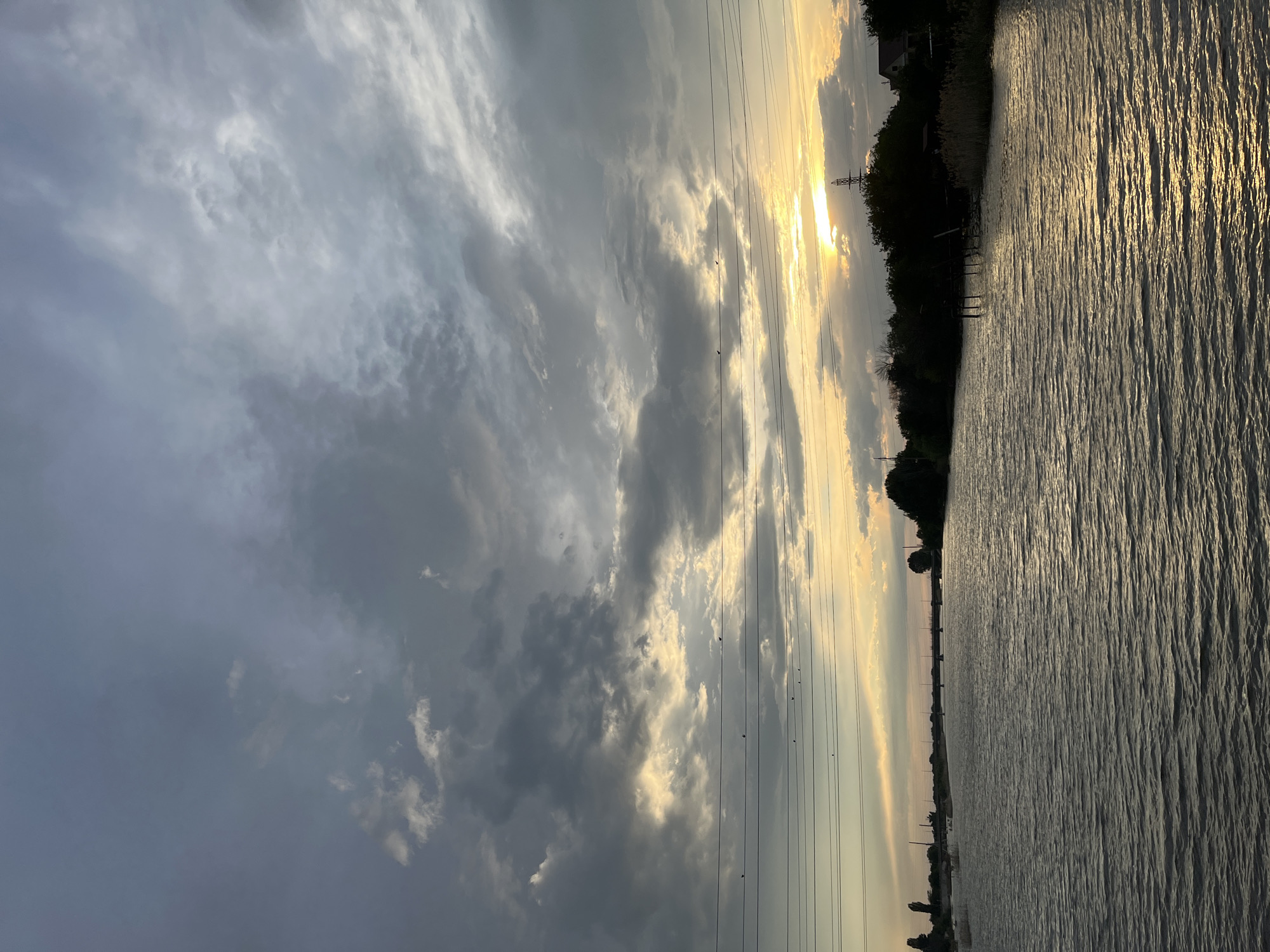 The image depicts a tranquil river scene at dusk in Krasnyye Barrikady, Russia. The sky is filled with dramatic, cloud-streaked twilight, with the setting sun casting a warm, golden glow through breaks in the clouds. Power lines with several small birds perched on them stretch across the middle ground of the image. The river's surface reflects the colors of the sky, creating a shimmering, textured effect. Along the far bank, a dense line of trees and vegetation is visible, with a bridge spanning the river in the distance. A few scattered structures, possibly houses, are discernible amongst the trees on the right bank. The overall impression is one of peaceful natural beauty as day transitions into night.