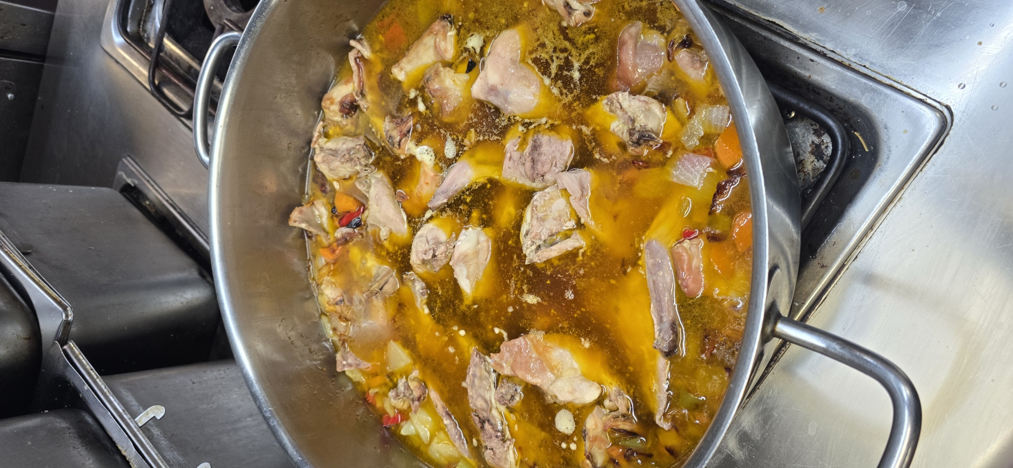 A close-up, high-angle shot shows a large pot of what appears to be a hearty stew or soup, filled with pieces of meat and vegetables, simmering on a commercial gas stovetop. The broth is a rich, golden-yellow color, with visible chunks of pale meat, diced orange carrots, and possibly white onions and red peppers. The metal pot has two sturdy handles and sits on a stainless steel surface that shows signs of use, with some minor staining and grease marks around the stovetop burners. The background is dominated by stainless steel surfaces and containers, typical of a professional kitchen environment. The scene suggests a food preparation setting, likely in a restaurant or commercial kitchen. No people are visible in the image. The location is identified as Sallent de Gállego, Spain.
