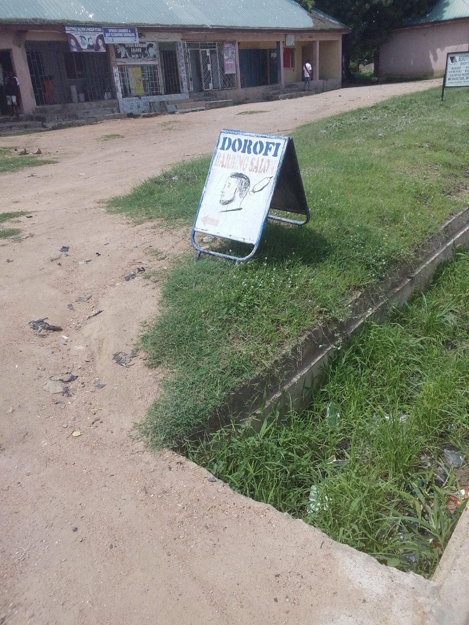 This is a medium-shot, eye-level, outdoor photograph of a street scene in Jalingo, Nigeria. The image is dominated by a dirt road and a grassy embankment with a concrete drainage channel running along its edge. In the foreground, an A-frame sign with the text "DOROFI BARBING SALOON" and a cartoon drawing of a man's head is propped up on the grass. The sign has a blue border and an arrow pointing to the left.
In the background, a row of shops lines the opposite side of the road. The shops have open fronts with bars and some display signage. One sign reads "BEAUTYMAX SALOON & MAKEUP STUDIO" and another "SPACO LAUNDRY & DRY CLEANING SERVICES". A person in white and red clothing can be seen walking near one of the buildings. The overall scene suggests a commercial area in a town.
The lighting indicates it is daytime, possibly late morning or early afternoon, with bright sunlight casting shadows. The weather appears to be fair. The presence of the unpaved road, the style of the buildings, and the visible signage suggest a developing urban environment. The grassy area and drainage channel indicate a somewhat neglected or utilitarian infrastructure. The image captures a typical street view with commercial establishments and a roadside advertisement.