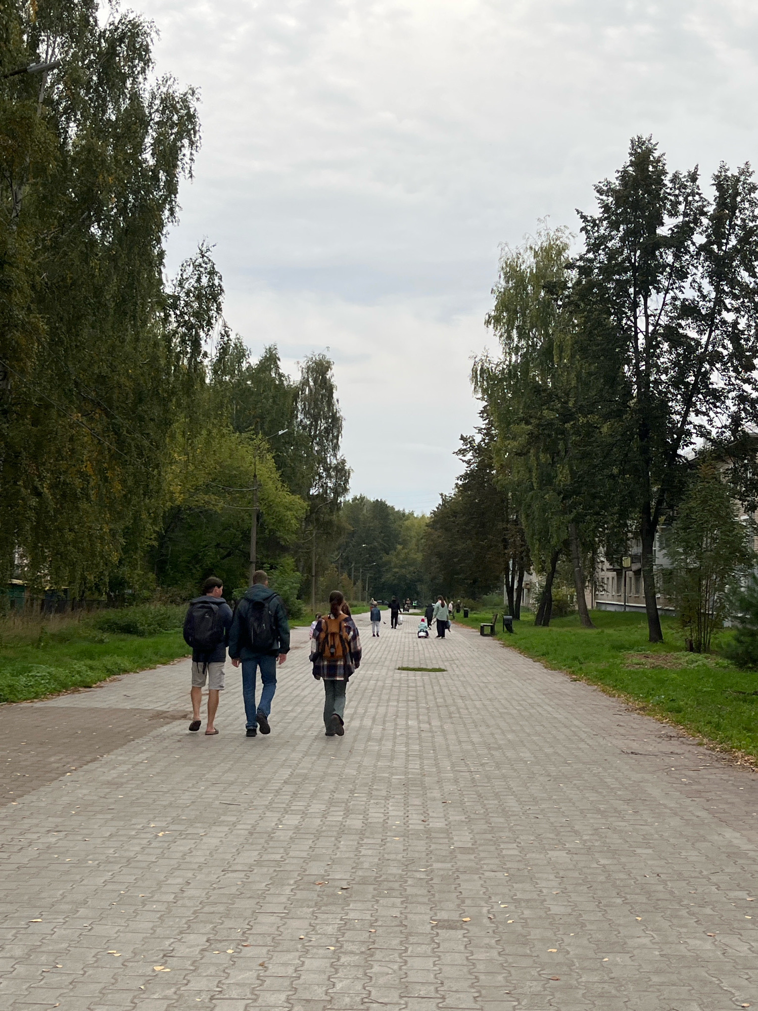 A paved walkway lined with trees runs through a park in Chaikovsky, Russia, under a cloudy sky. Three individuals walk away from the camera on the left side of the walkway. The first person, on the far left, wears shorts and sandals. The second person, in the middle, wears jeans and a backpack. The third person, on the right, wears a plaid shirt and a backpack. Further down the walkway, other individuals are visible, some with a child and a stroller. A grassy area with benches and trees borders the right side of the walkway, with apartment buildings in the background.
