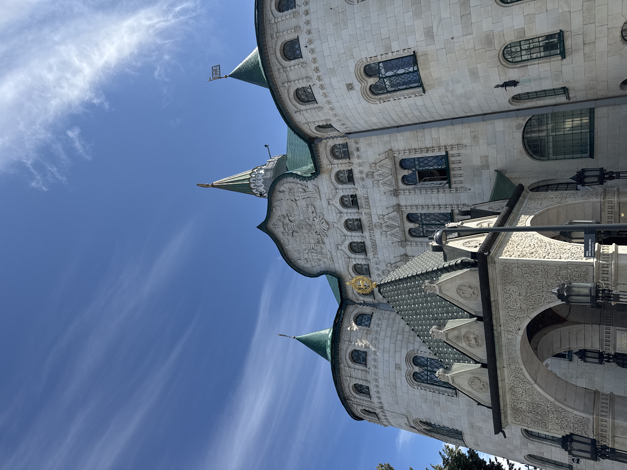 The image shows a low-angle shot of the Nizhny Novgorod State Bank building in Nizhny Novgorod, Russia. The building is made of light-colored stone and features intricate architectural details, including multiple turrets with green roofs, arched windows, and decorative carvings. A prominent golden emblem adorns the roof of a central section. The sky above is a clear blue with wispy white clouds. The scene appears to be captured on a bright, sunny day, with no people or vehicles visible. The overall impression is one of grandeur and historical significance.