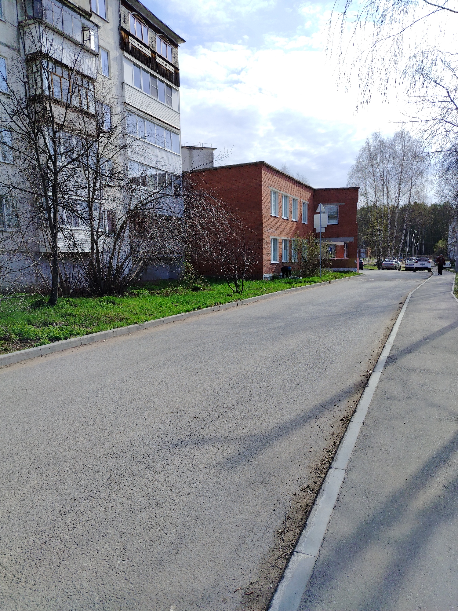 The image shows a street scene in Noviy, Russia. A gray, multi-story residential building is on the left. Next to it, a one-story brick building with white-framed windows. A paved road runs along the front of the buildings, with a sidewalk on the right side. Several parked cars are visible further down the street, and a person is walking away from the camera on the sidewalk. A small patch of green grass separates the road from the building on the left. Bare trees stand in front of both buildings. The sky is cloudy.