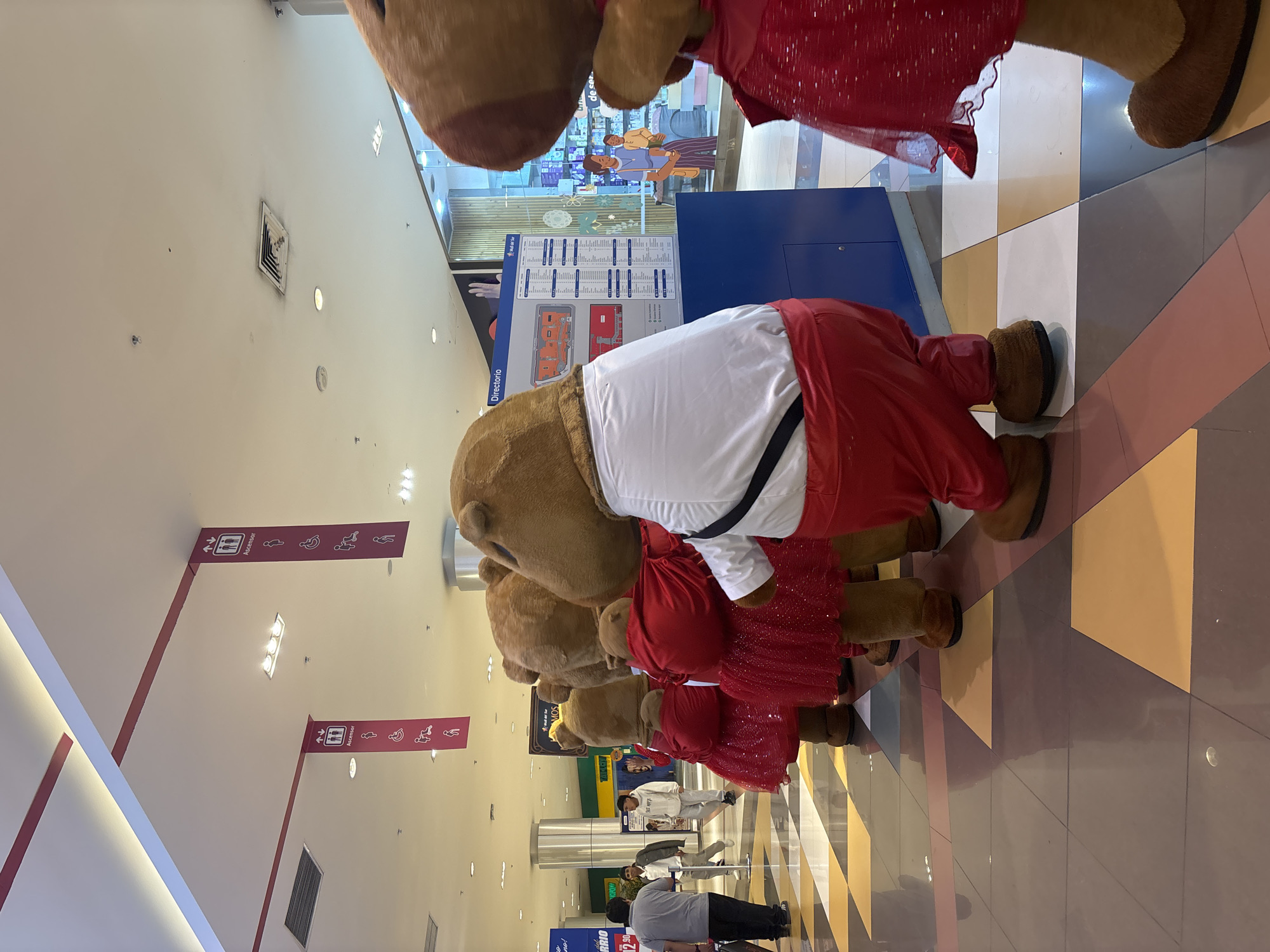 A photograph shows a row of costumed characters in what appears to be a shopping mall. The characters, presumably bears, are wearing red and white outfits. The mall's directory sign is visible behind them, along with general store information and a directory map. People are walking in the background. Overhead signs indicate the location of elevators ("Ascensor"), and the floor is tiled in a geometric pattern of brown, white, and yellow. The specific location within San Juan de Miraflores, Peru, cannot be determined solely from the image.
