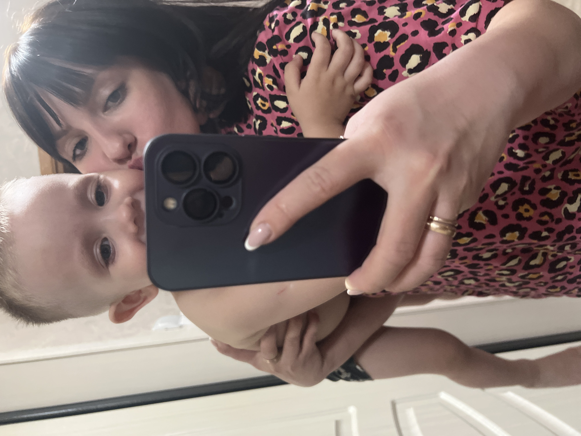 A selfie photograph shows a woman and a baby, seemingly a mother and child. The woman has dark hair and is wearing a pink top with a black and yellow leopard print. She is kissing the baby's cheek while holding a smartphone, likely taking the photo. The baby has fair skin and is looking towards the camera. The background is a light-colored wall or door with white paneling. The photo appears to have been taken indoors in Марковский, Russia.