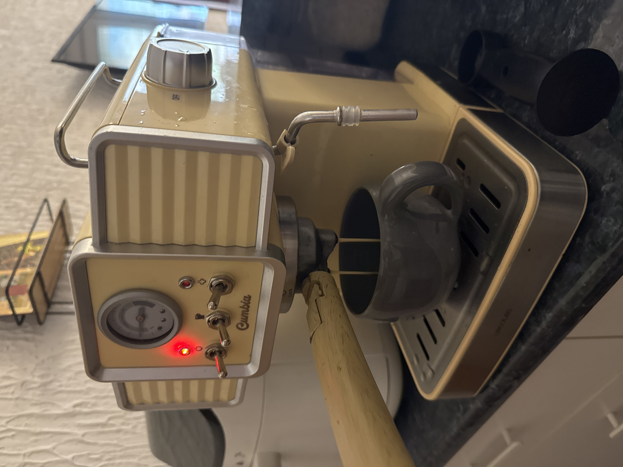 The image displays a retro-styled coffee machine, predominantly cream or pale yellow with silver accents, positioned on what appears to be a dark, speckled kitchen countertop. The machine is oriented vertically in the photograph.
The main subject is the coffee machine, which features a prominent front panel with a pressure gauge, three toggle switches, and a red indicator light that is illuminated, suggesting the machine is powered on or heating. Below the gauge, the brand name "Cumbia" is visible. A rotary knob on the top right surface of the machine has "OFF" inscribed on it. Small water droplets are scattered across the top surface.
To the right, a silver steam wand with a clear rubber ring extends from the machine. Below the steam wand, a dark grey coffee mug sits on a rectangular, silver-rimmed drip tray with slotted perforations. A wooden handle, possibly belonging to a portafilter, is inserted into the group head of the machine, with "Lock" visible next to the attachment point. This wooden handle appears somewhat worn.
In the background, behind the machine, dark, cylindrical objects are visible, possibly coffee accessories like a tamper or grinder. Part of a metal rack can be seen in the blurry background on the left. Faint, partially obscured text, possibly "CMK4300", is visible on the silver rim of the drip tray. The overall setting is indoors, likely a kitchen. No people are visible, and the time of day or weather cannot be determined.