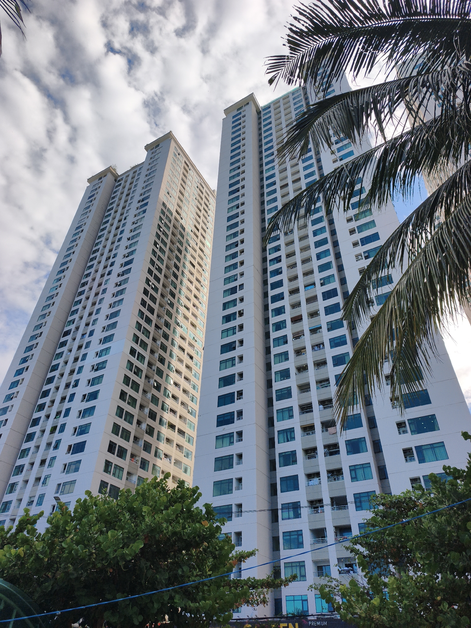 The image shows two tall, white apartment buildings with many windows, some of which are tinted blue. Palm fronds are in the foreground, obscuring parts of the buildings. Lush green trees are visible at the base of the buildings. The sky is partly cloudy. The architectural style suggests a modern residential complex in a tropical or subtropical urban setting, consistent with locations like Nha Trang, Vietnam, known for its coastal high-rises and palm trees. There are no people or visible activity depicted in the scene.