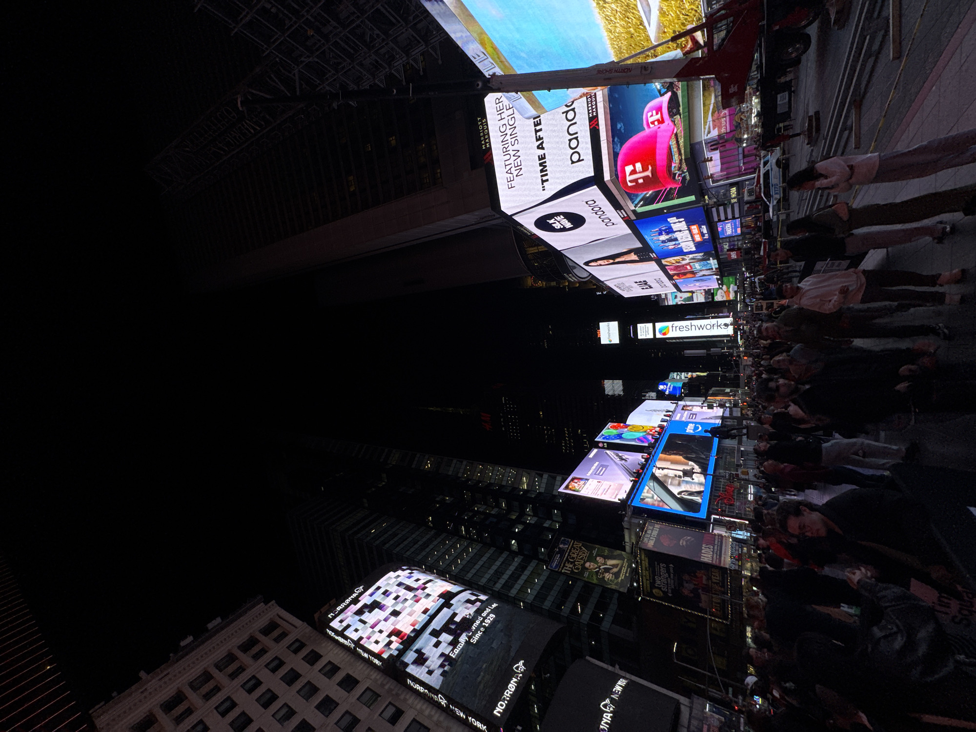 This image captures a vibrant nighttime scene in Times Square, New York City. Several large, illuminated digital billboards dominate the view, advertising various products and shows, including Pandora jewelry, the musical "The Great Gatsby," and a campaign for "Time After Time". People are densely packed together, walking and looking at the displays. Some are partially obscured by the camera's angle. Modern skyscrapers surround the open space, and some buildings are partially dark against the night sky. The image is taken from a tilted perspective, giving a dynamic, almost disorienting feel to the urban scene.