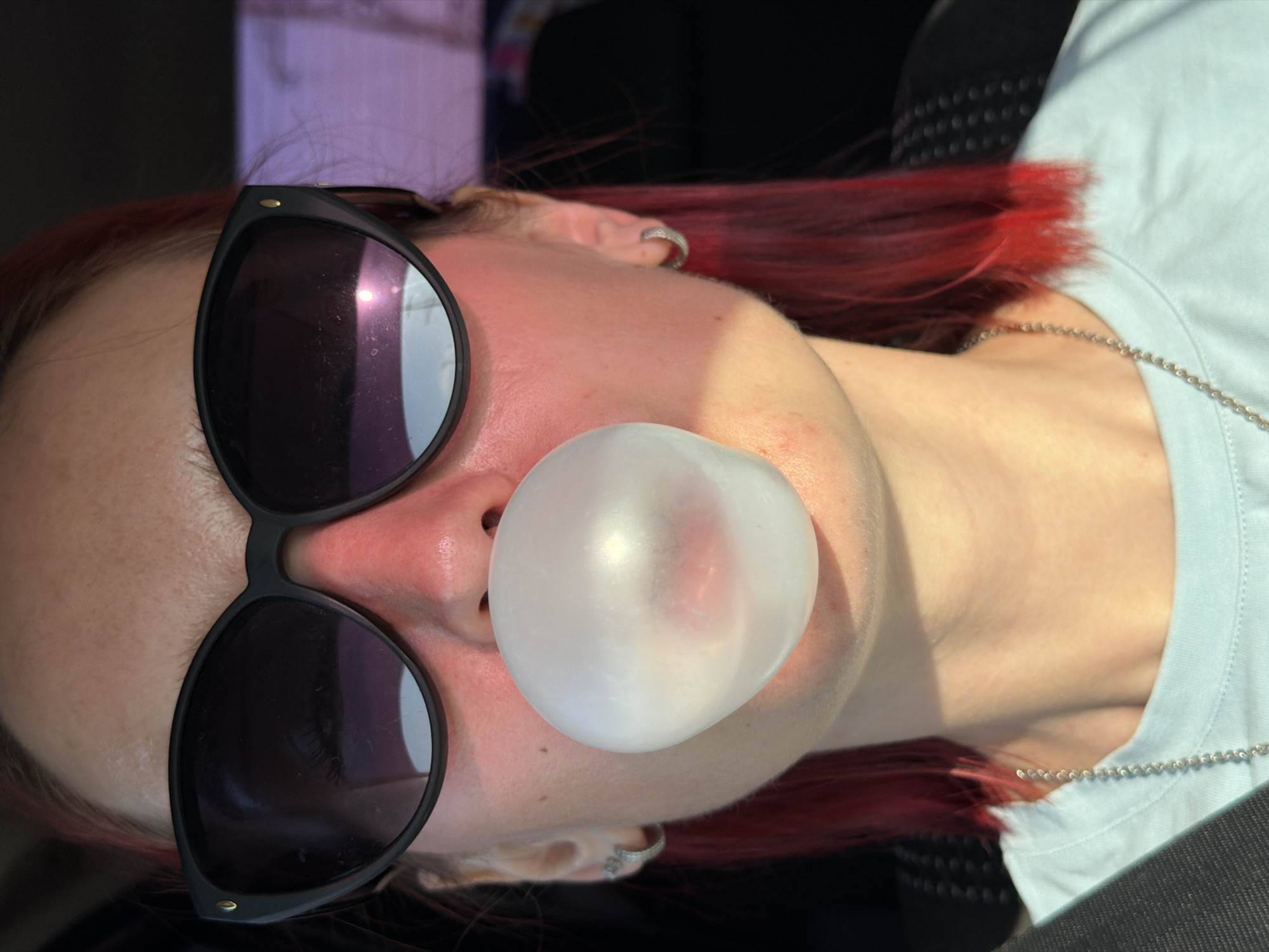 A woman with red hair is blowing a bubble. She is wearing sunglasses and a light blue shirt with a silver chain. She also has ear piercings. The location cannot be verified based on the image.