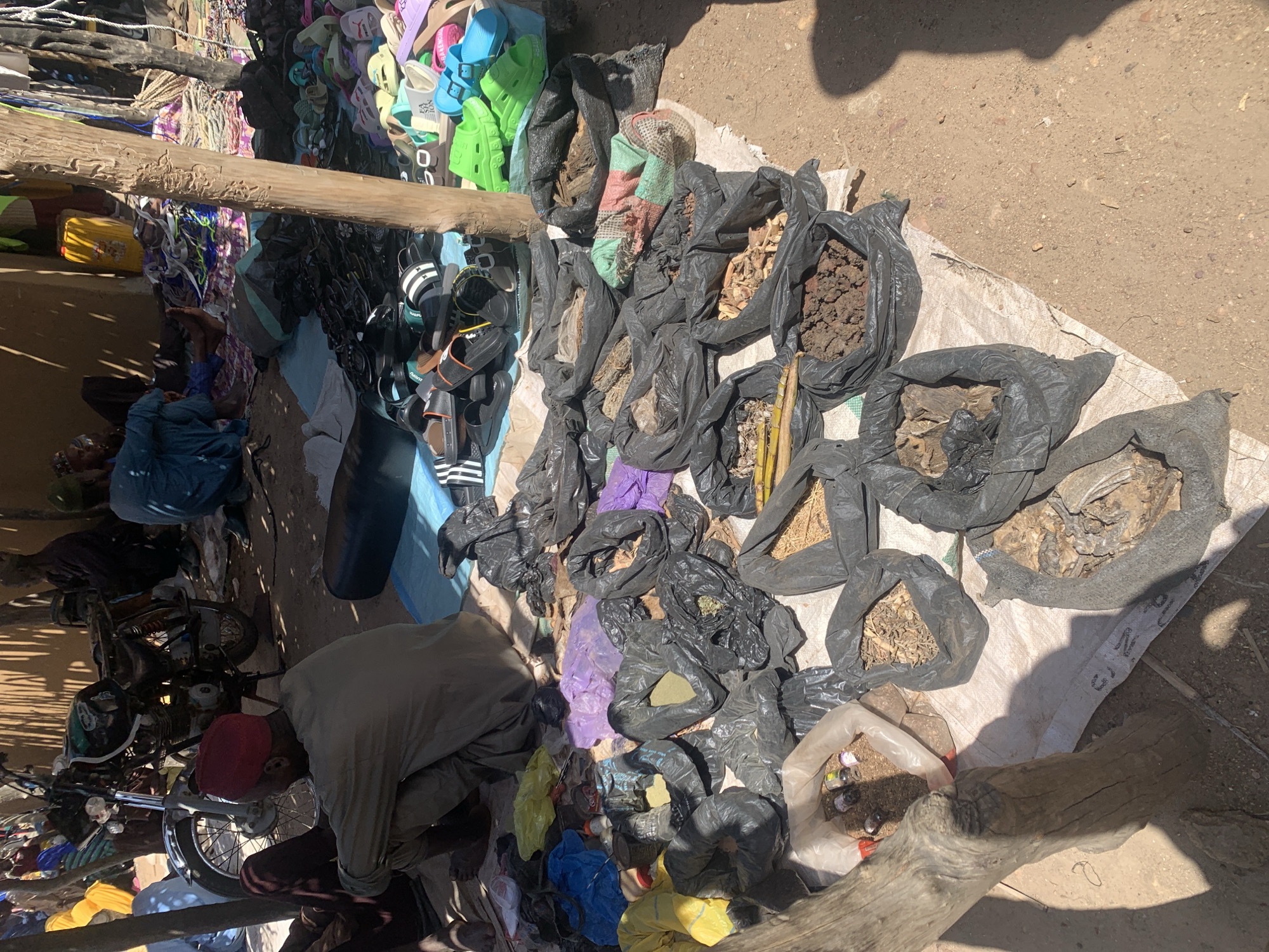 This media file captures a vibrant outdoor market scene, likely in Dull A, Nigeria. The primary subjects are people and a variety of goods displayed for sale. The setting is a dusty, sun-drenched area, suggesting a warm, possibly dry climate and a daytime time of day, indicated by the strong shadows cast by overhead objects and people.
Various objects are prominent, including a large assortment of colorful flip-flops and sandals neatly hung or displayed, suggesting a footwear vendor. Beside these, an array of black plastic bags filled with what appears to be dried goods, soil, roots, and possibly medicinal herbs are laid out on a white tarp. Some of these bags are partially open, revealing their contents. Other items include a motorcycle, suggesting transportation or goods delivery, and various pieces of clothing or textiles, some of which are brightly colored, like purple and yellow bags, and others appear to be sacks. A large piece of driftwood is positioned across the foreground.
The activity taking place is clearly a commercial transaction, with vendors displaying their wares and potential customers present, although the focus is more on the goods than the interactions. The overall impression is one of a bustling marketplace, with a sense of daily commerce. The strong sunlight and shadows imply it's a clear day. The general atmosphere seems lively and active, typical of a market. There is no visible text in the image.
