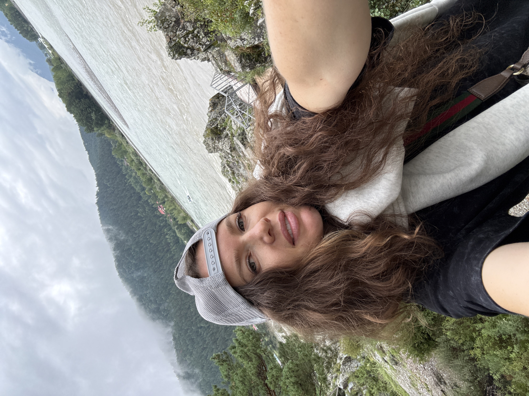 A woman wearing a gray baseball cap and a white scarf takes a selfie outdoors. She has long, curly brown hair and is smiling at the camera. The background shows a wide, light-colored river and a tree-covered mountain range under a cloudy sky. The location is likely Elekmonar, Russia, given the scenery. The woman is wearing a black shirt and appears to be on a rocky incline near the river, with some greenery and a metal staircase visible.