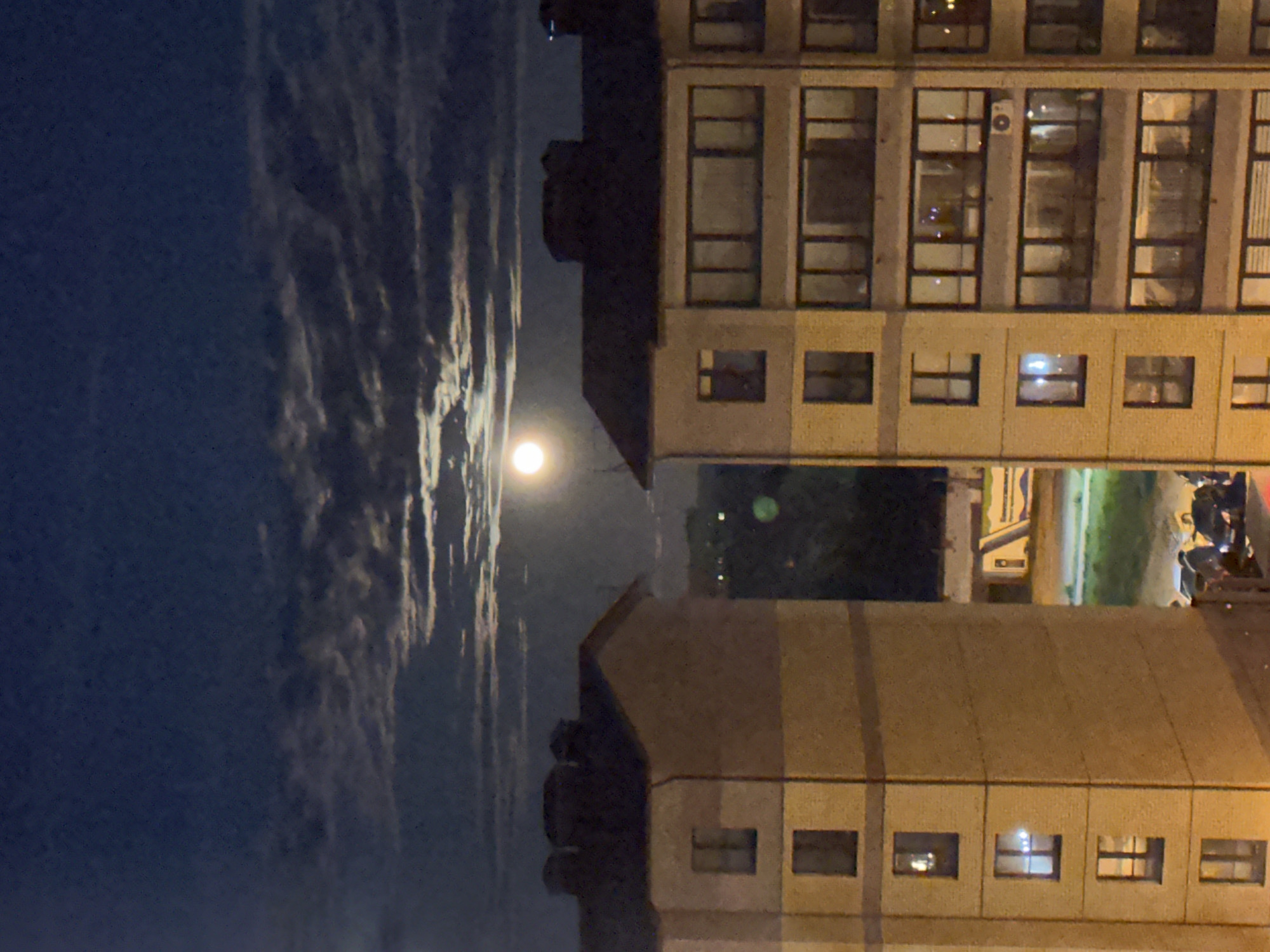 Captured at night, the image reveals a cityscape of Vladimir, Russia. The composition is oriented vertically, with the night sky and moon on the left and the urban buildings occupying the right. The sky presents a mix of dark blue interspersed with cloudy formations. The moon shines prominently, positioned between the buildings. The buildings themselves are constructed with tan or light-colored concrete and feature rows of windows, some illuminated. The scene is illuminated by both moonlight and artificial light sources emanating from the buildings and surrounding areas. The overall impression is that of a quiet, urban nightscape.