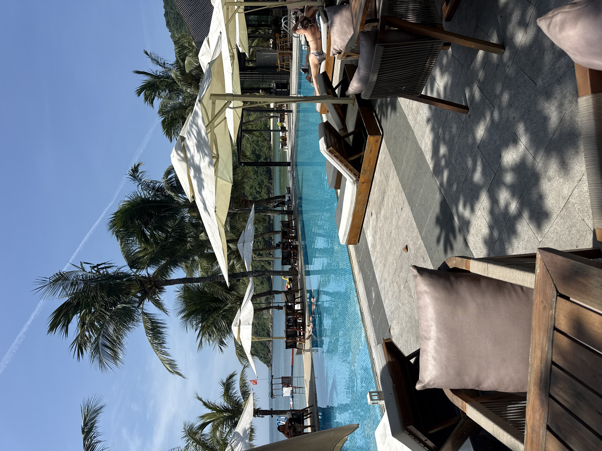 The image shows a poolside scene in an unknown city. A swimming pool runs vertically through the frame, with white umbrellas and palm trees lining its side. Lounge chairs with cushions are placed along the pool, with a few people relaxing on them. In the background, the ocean can be seen, and a contrail is visible in the sky. The foreground contains wooden chairs with cushions, and the ground is tiled. The sun casts shadows on the ground and furniture.