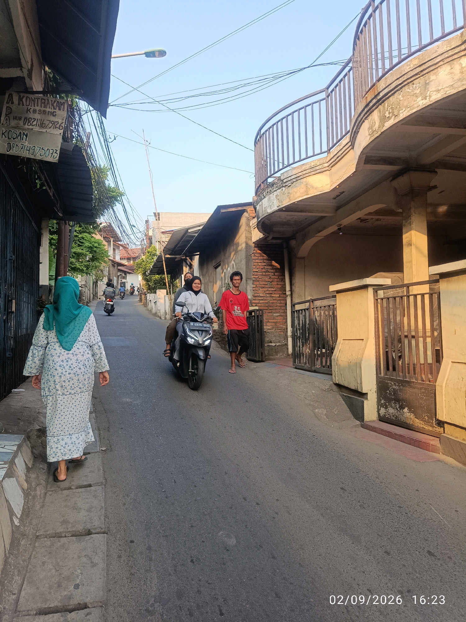 In an outdoor urban setting, a woman in a light-colored, patterned dress and a teal hijab walks away from the camera on the left side of a narrow street. Ahead of her, a motorcycle carrying two people, a driver and a passenger, travels down the street. The motorcycle has a license plate that reads "B 2878 SKG". To the right of the motorcycle, a man in a red t-shirt and black shorts walks towards the camera. Further down the street, several other motorcycles are visible. The buildings on either side of the street are typical of a residential area, with some appearing to be for rent, as indicated by a sign on the left that reads "A KONTRAKAN KOSONG" and displays phone numbers. The sky is clear and blue, and the sun casts long shadows, suggesting it is late afternoon. The street is paved with asphalt, and a concrete sidewalk is visible on the left. A modern balcony with a railing extends from a building on the right. The timestamp "02/09/2026 16:23" is displayed in the lower right corner of the image.