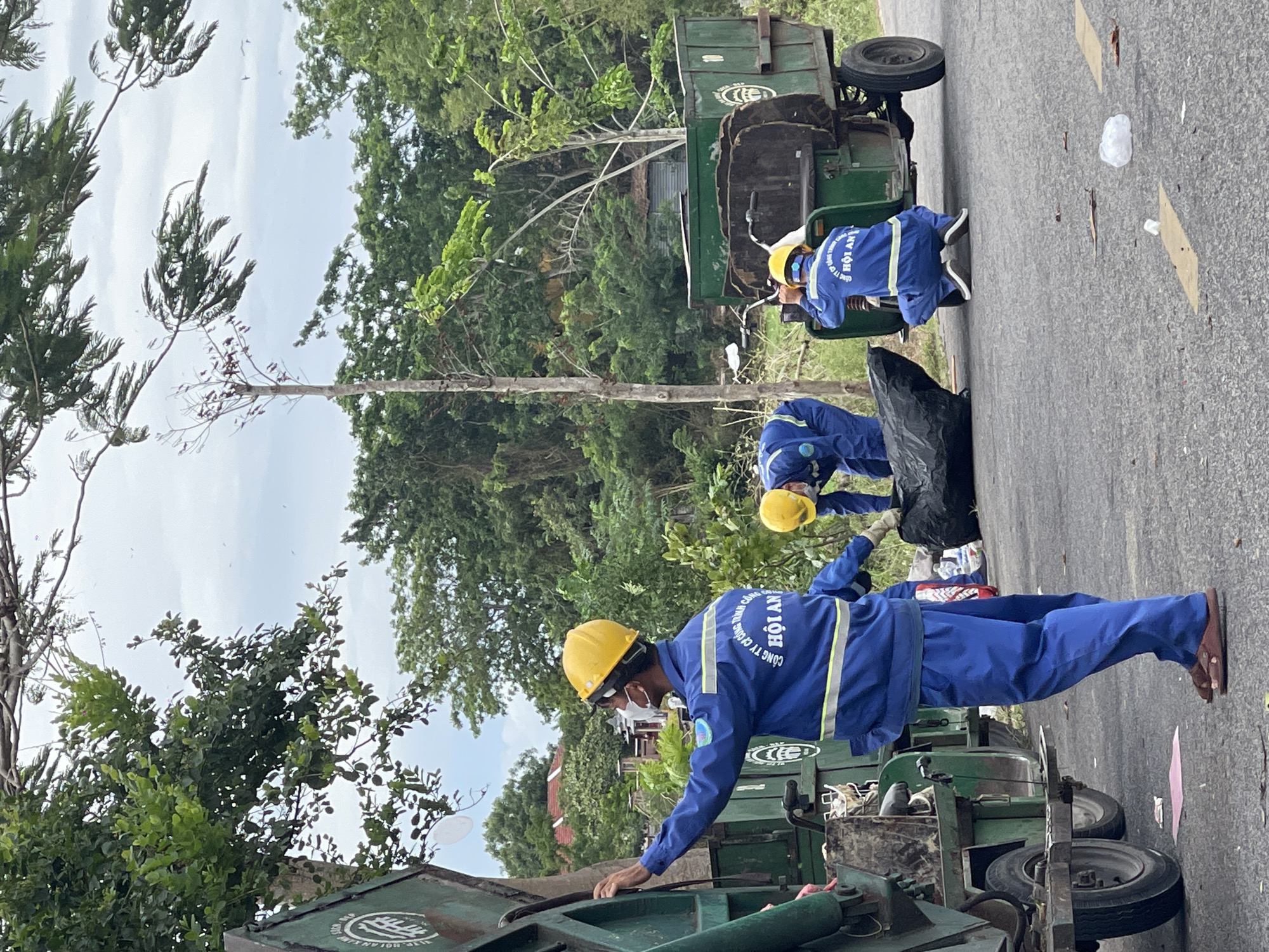This image shows three sanitation workers in blue uniforms and yellow hard hats working on a roadside in Hoi An, Vietnam. They are collecting trash, with one worker in the foreground standing by a garbage truck, another in the middle bending to pick up a large black trash bag, and a third worker in the background crouching near a smaller trash cart. The environment is semi-urban, with trees and vegetation lining the road, and the sky is overcast. The garbage trucks are green, and there is loose trash visible on the road. The text on the workers' uniforms reads "CÔNG TY TNHH MỘT THÀNH VIÊN MÔI TRƯỜNG ĐÔ THỊ HỘI AN," which translates to "Hoi An Urban Environment One Member Limited Liability Company."