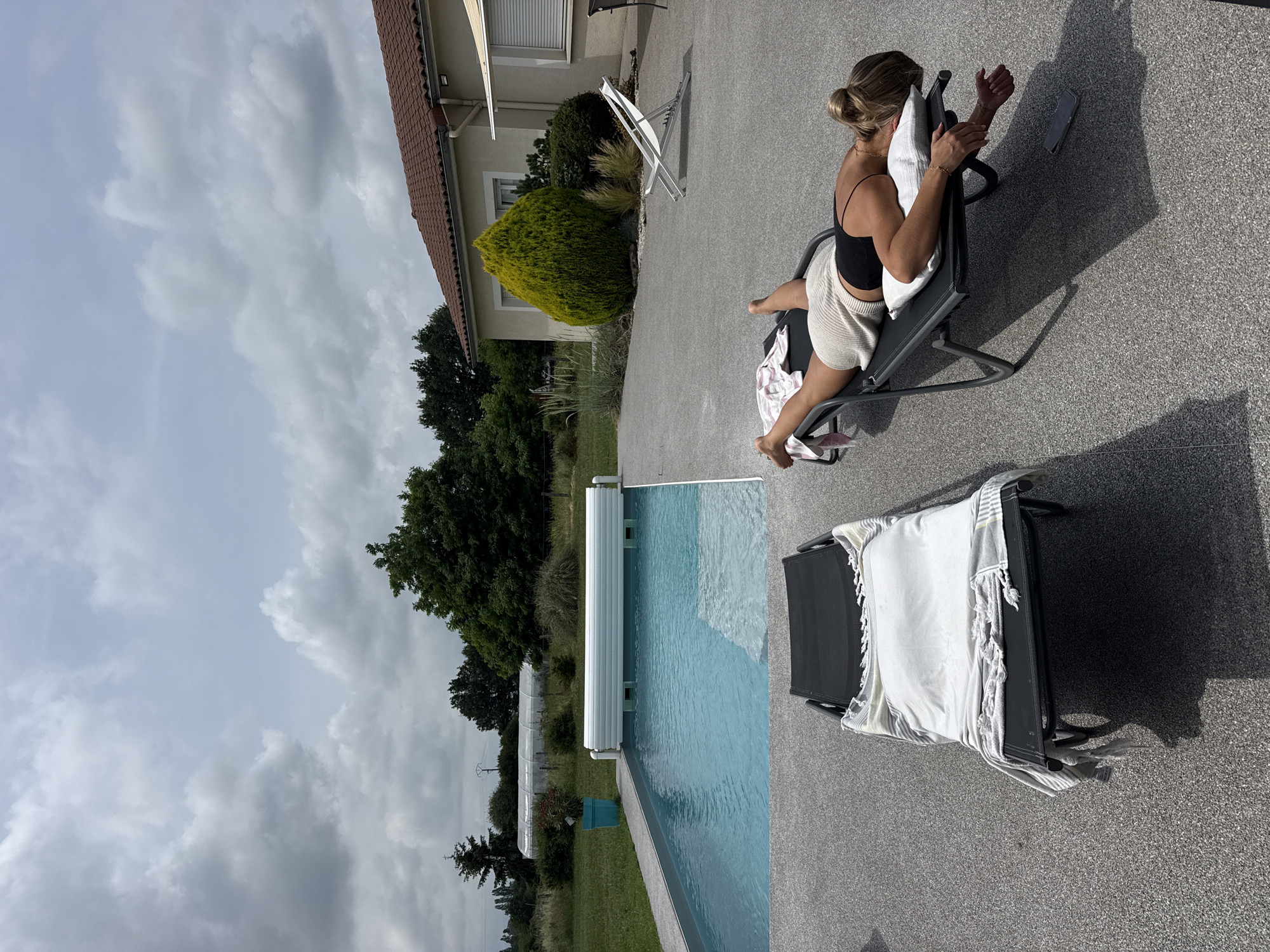 The image shows a scene in Veauchette, France. A woman is seated in a lounge chair on a concrete patio. She has a pillow behind her and is looking at a smartphone in her hands. Next to the chair is a pool with a retractable cover. Another lounge chair with a white blanket is next to the pool. In the background is a house with a red tile roof, a manicured bush, trees, and a cloudy sky.