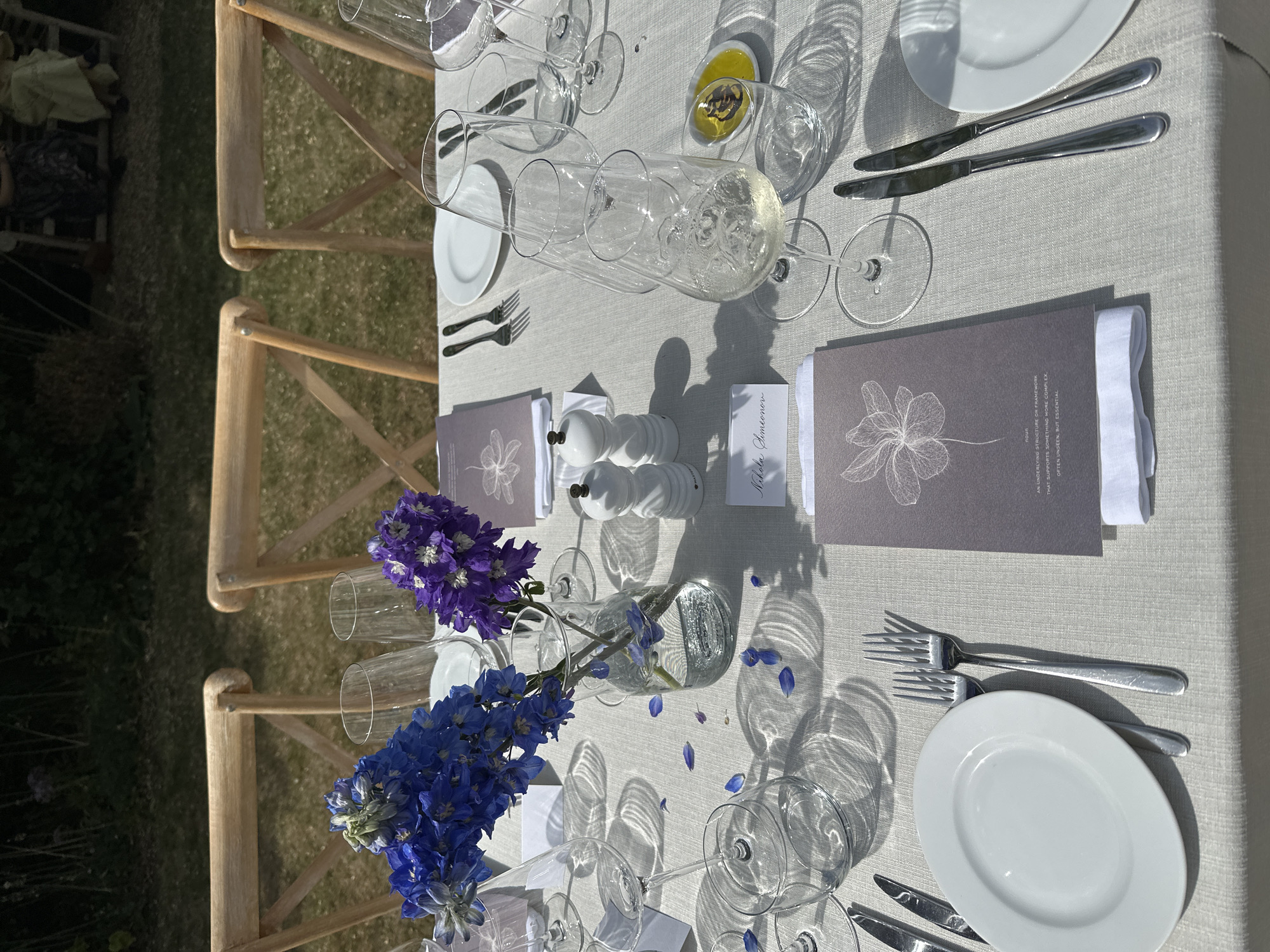 The image depicts a set table outdoors, likely at a wedding or formal event. The tablecloth is light grey, and the place settings include white plates, silver cutlery, multiple wine glasses, and menus with a floral design. Each setting also has a small name card and a salt and pepper shaker. A glass vase with blue and purple flowers serves as a centerpiece. The chairs are light wood with a cross-back design. The location appears to be outdoors on a grassy area. It's impossible to confirm the location as Cambridge from the image alone.