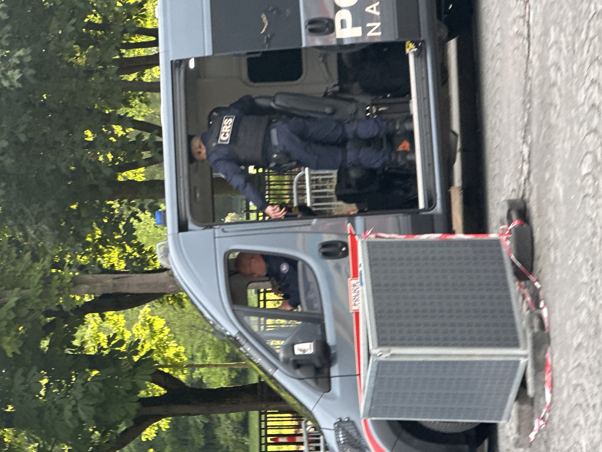 A gray police van with a person visible in the driver's seat and a second person in uniform standing in the back with "CRS" clearly displayed on the back of the vest. The vehicle is parked on the side of the street. The location appears to be Paris, France.