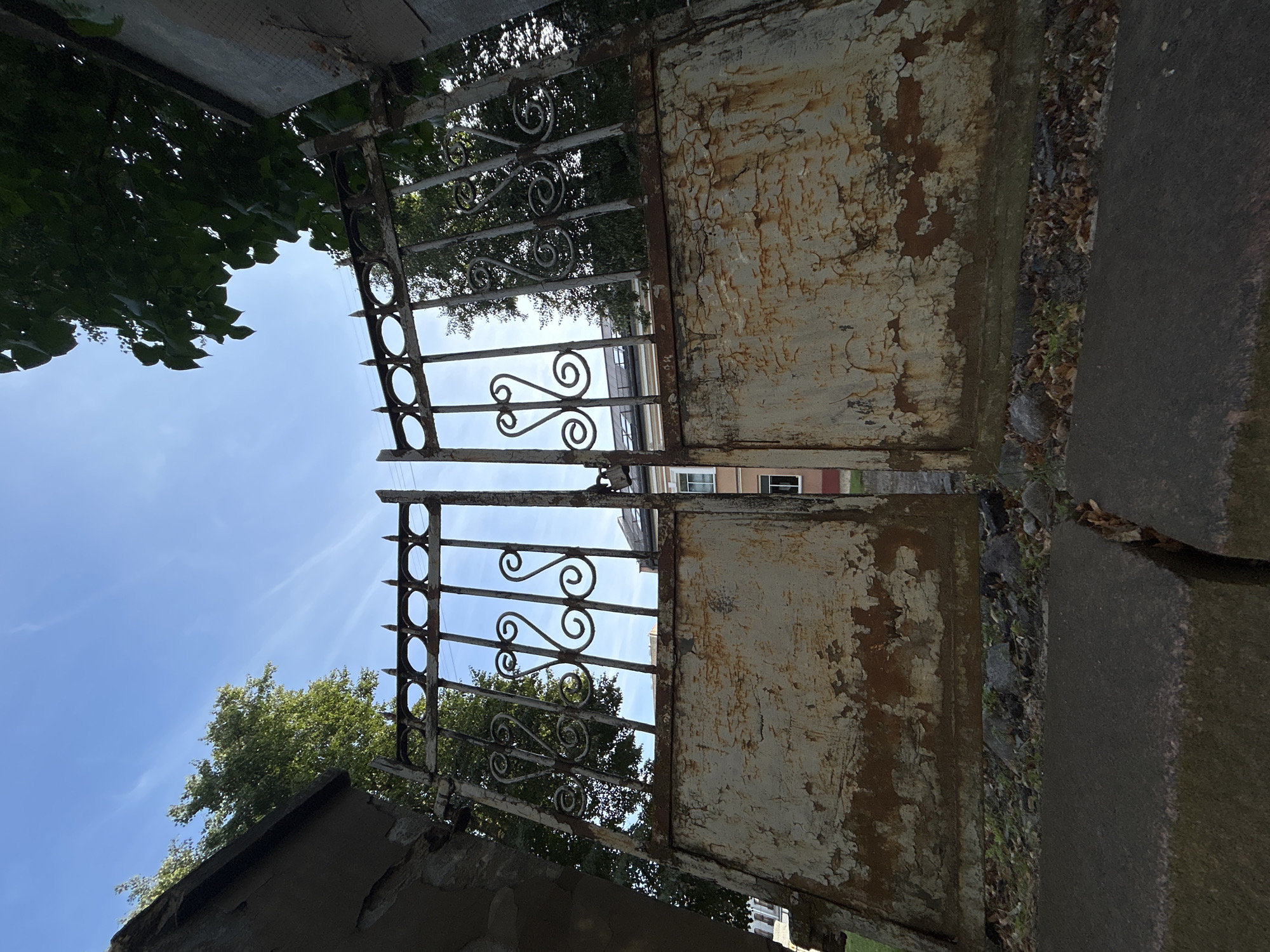 This is a low-angle, wide shot of an old, rusty, double-leaf metal gate in Vilnius, Lithuania, possibly in the Šnipiškės district, based on the architectural style of the building visible through the gate. The gate is made of wrought iron with decorative scrollwork and pointed finials. The lower panels of the gate are heavily weathered, with peeling paint and visible rust. The gate is open, revealing a glimpse of a building with several windows and a tiled roof behind it. Trees with green foliage are visible on either side of the gate and peeking through the top. The sky above is a clear blue with a few wispy white clouds. In the foreground, a cracked concrete surface is visible. The overall impression is one of age and disrepair, contrasting with the bright, clear sky.