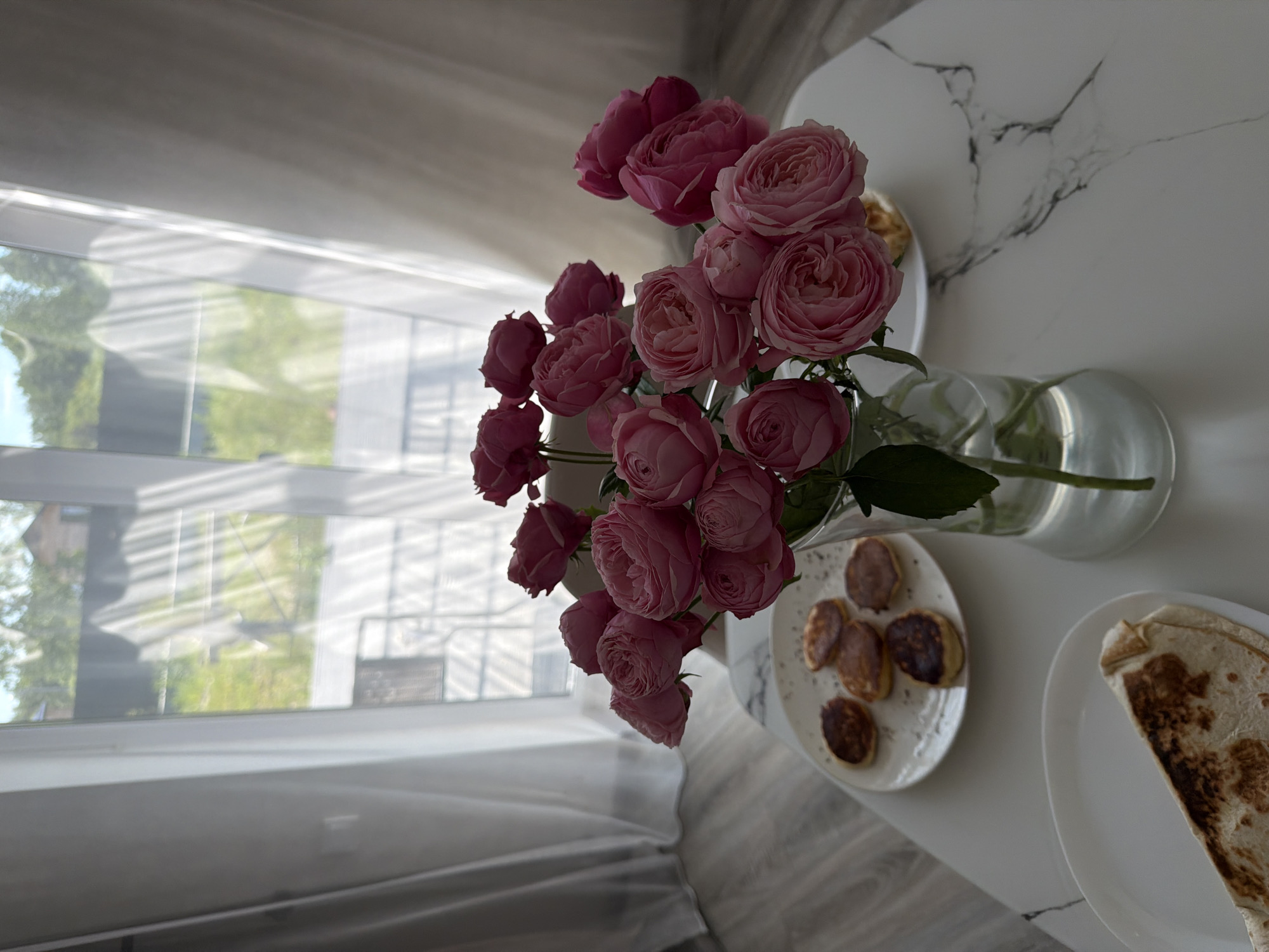 A still life photograph features a bouquet of pink roses in a clear glass vase on a white marble-patterned table. To the left of the vase, a white plate holds several small, round pancakes. Another white plate below it contains a folded flatbread wrap. In the background, sheer white curtains are drawn, revealing a blurred view of greenery and a fence outside, suggesting a residential area in Malinovka, Russia. The lighting indicates it is daytime. There are no people visible in the image.