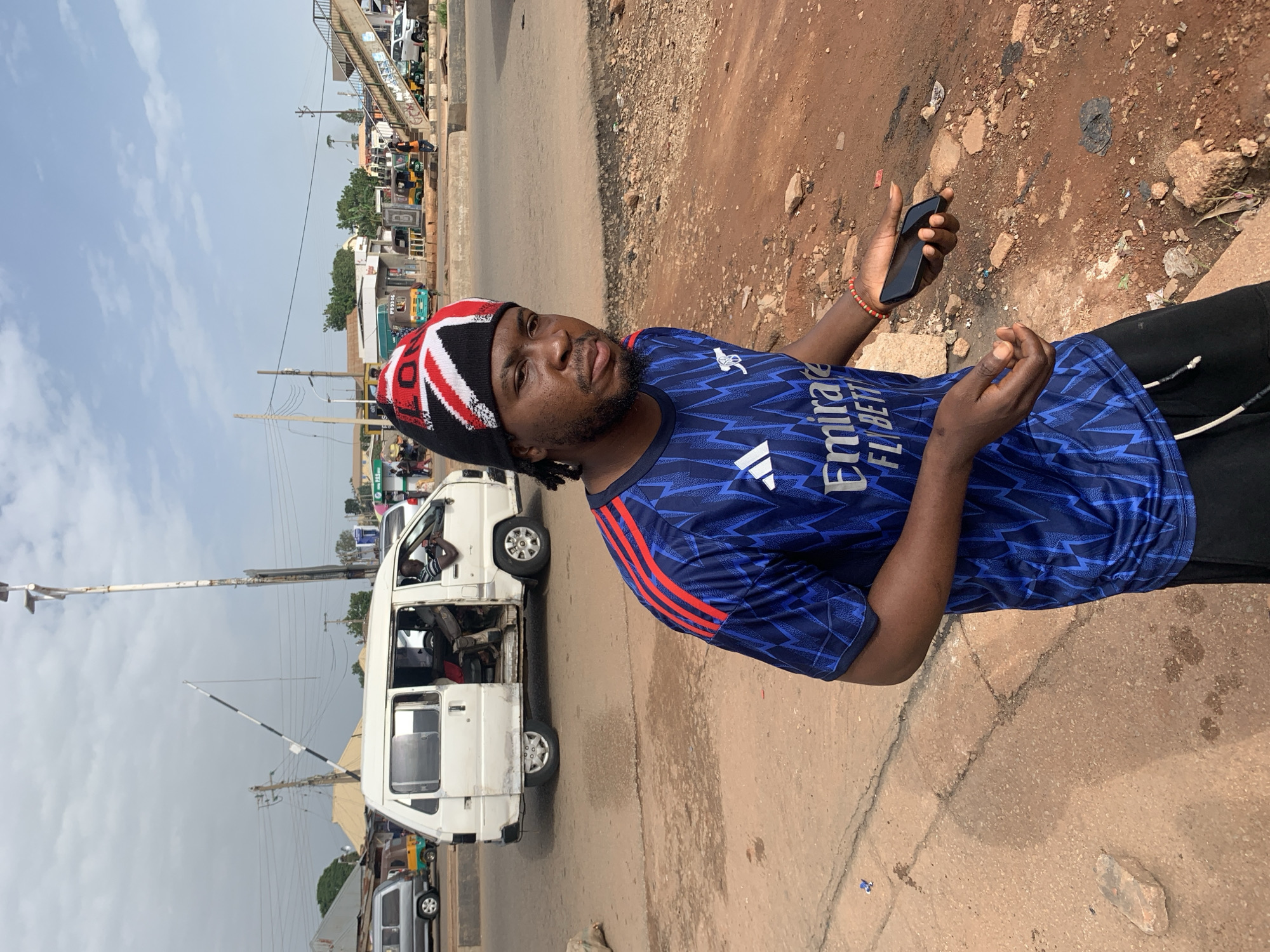 A young Nigerian man stands in a dusty, outdoor setting, likely in Gura Top, Nigeria. He is wearing a blue and red patterned soccer jersey with the logo "Emirates Fly Better" visible. On his head, he wears a black, white, and red knitted beanie adorned with a Union Jack pattern. He holds a black smartphone in his right hand, which is outstretched, and his left hand is also extended, palm up. The background features a white van with its doors open, a traffic light signal, and several other vehicles, including tuk-tuks and a white SUV, suggesting an urban or semi-urban environment with moderate traffic. The ground is covered in reddish-brown dirt and scattered debris. The sky is overcast with some blue patches.