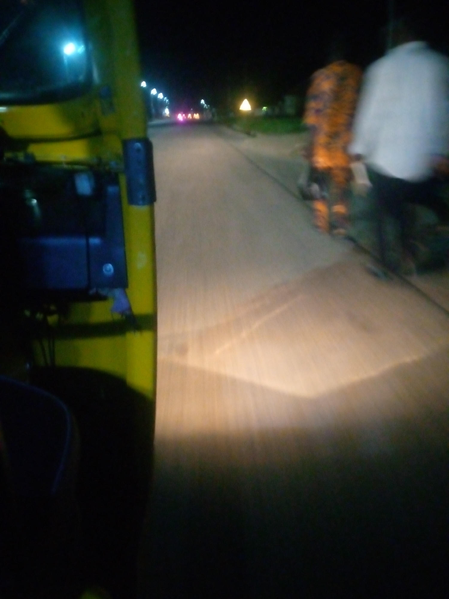 The media file is a nighttime photograph taken from inside a yellow vehicle, likely a tuk-tuk, as indicated by the visible yellow bodywork and dark interior in the foreground. The scene depicts a street at night, with several streetlights visible along the left side of the road, casting a blueish glow.
In the mid-ground, two figures are walking away from the camera. The person on the right is wearing a white garment, and the person to their left is wearing a brightly colored orange and black patterned outfit. The motion blur suggests the vehicle is moving, and the figures are also in motion.
The road surface is visible and illuminated by the headlights of the vehicle, creating bright streaks of light. In the distance, there are more blurred lights, possibly from other vehicles or buildings. A triangular illuminated object can be seen in the far distance, potentially a sign or a building feature. The overall atmosphere is dark, typical of nighttime. No text is visible in the image. The setting appears to be a street in an urban or semi-urban area.