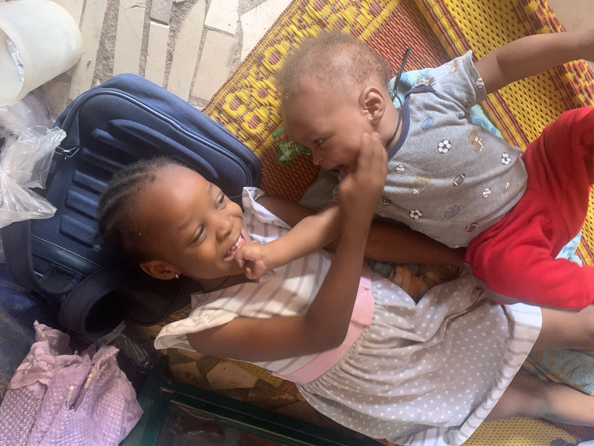 In a brightly lit indoor space in Sabon Tasha, Nigeria, a young girl and a baby are lying on a woven mat. The girl, with braided hair and a small earring, is wearing a striped and polka-dot dress with a pink sash. She is smiling and touching the baby's face. The baby, with short, light brown hair, is wearing a gray shirt with sports-themed prints and red pants. They appear to be interacting playfully. Next to the girl is a dark blue backpack, and partially visible nearby are plastic bags and a white container. The floor is a mix of tiled and concrete surfaces.