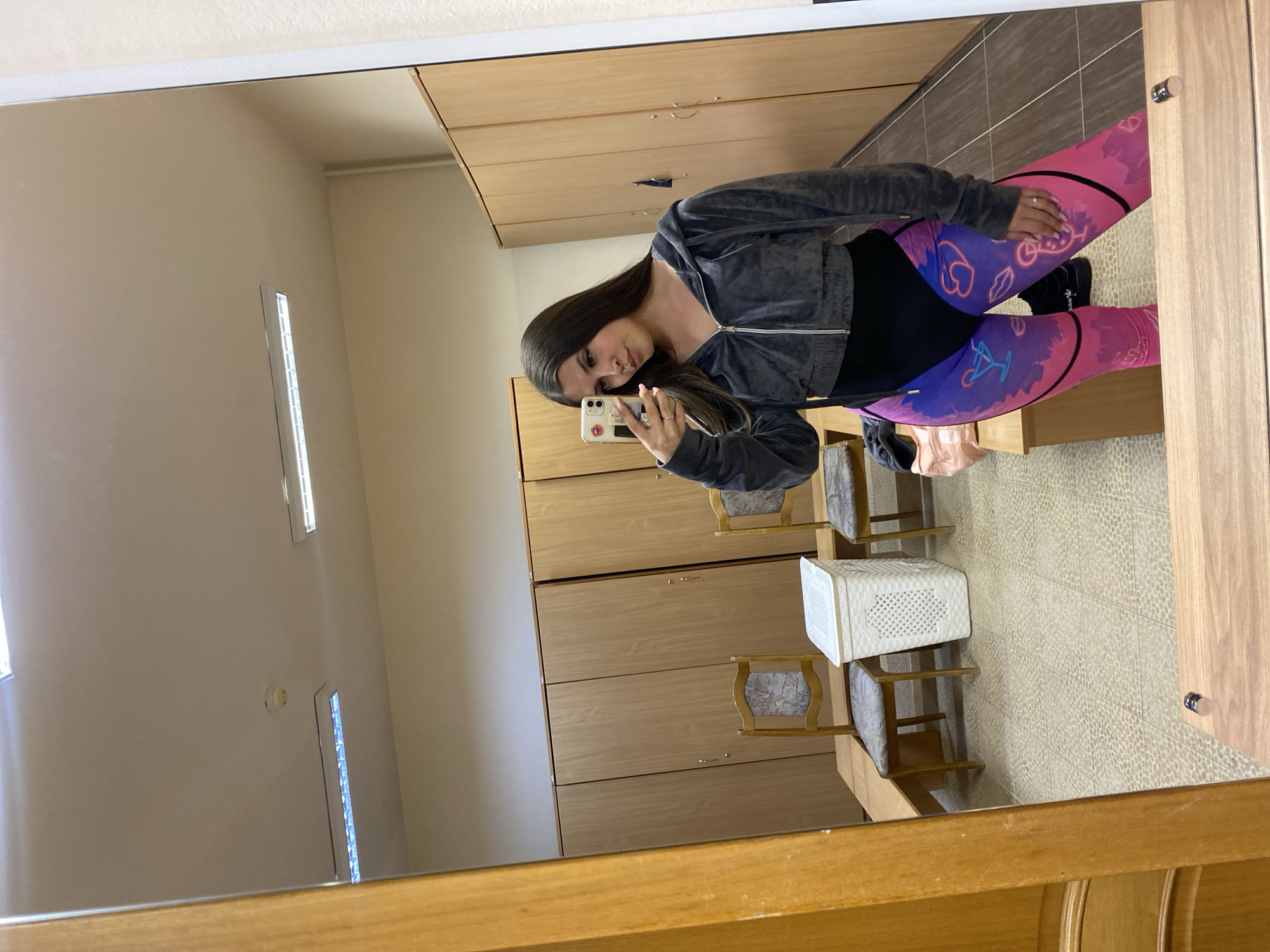 A young woman is captured in a mirror selfie within an indoor setting, likely a changing room or locker area in Chaikovsky, Russia. She stands centered, holding a white iPhone with a light-colored case and partially visible text ("hello...") in her left hand, which is raised to take the photo. She has long, dark hair and is looking directly into the phone/mirror, showing a calm expression.
She is dressed in a dark grey, velvety cropped zip-up hoodie worn over a black top, paired with vibrant, multi-colored leggings. The leggings feature a base of pink and purple, adorned with graphic symbols such as hearts, lips, a bicycle, and a figure on a swing. A silver ring is visible on her right ring finger.
The background, reflected in the mirror, shows a functional space. Along the upper and left sides of the reflection are rows of light wooden lockers or wardrobes with silver handles. Two wooden chairs with light patterned upholstery are visible, along with a white, rectangular, perforated plastic basket on the floor. The floor is covered with light-colored, speckled tiles. The walls are plain and light-toned, with fluorescent strip lights providing illumination from the ceiling and a wall. A fire alarm or smoke detector is also visible on one of the walls. The mirror itself is framed by a light wooden border.