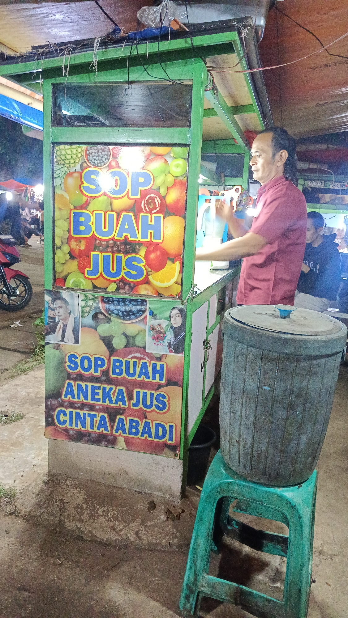 In Tajurhalang, Indonesia, a street-side fruit juice stand displays a vibrant sign featuring various fruits and the text "SOP BUAH ANBKA JUS CINTA ABADI". A man in a maroon shirt is actively preparing a drink behind the counter, with a large blue plastic bin on a green stool nearby. The scene appears to be at night, with artificial lighting illuminating the area and several other people visible in the background, some under tents. A red motorcycle is parked on the left side of the image. The ground is paved with dirt and concrete.