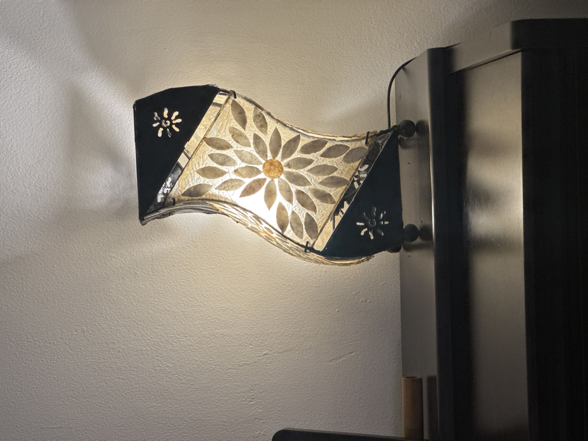 A wall-mounted lamp is illuminated against a textured, light-colored wall. The lamp has a curved shape with a floral design made of leaves on a translucent material. Black, angular metalwork frames the sides of the lamp, featuring cut-out floral patterns. A dark grey, modern cabinet is visible on the right side of the frame, attached to which the lamp is mounted. A dark electrical cord extends from the lamp. The lighting creates shadows and highlights on the wall. The location is not discernible from the image.