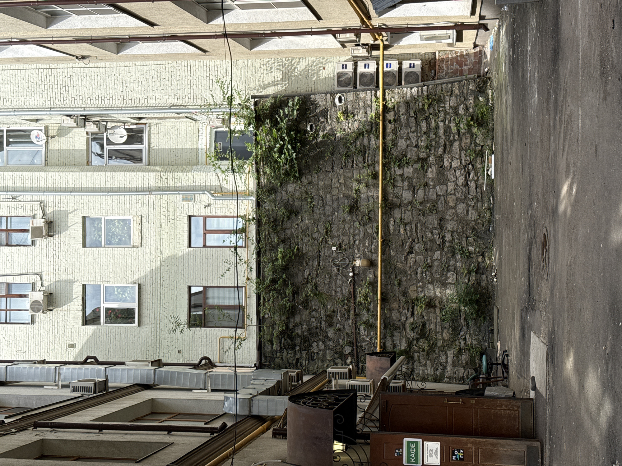 The image depicts a narrow courtyard between two buildings in Moscow, Russia. The left building is clad in grey material with several windows and prominent external ventilation ducts. A brown wooden door with a green sign that reads "КАФЕ" is visible on the ground floor. To the right of the door, there is a green office chair and some debris.
The right building is constructed of light-colored brick and also features numerous windows, air conditioning units, and satellite dishes. A rough stone wall, covered in vines and plants, dominates the central portion of the image, acting as a boundary or retaining wall. A bright yellow pipe runs horizontally across this stone wall. The ground is paved with asphalt, showing signs of wear and tear, with some patches of concrete and scattered debris. The scene appears to be an urban service area or a less-maintained courtyard, likely in a historical district given the architecture.