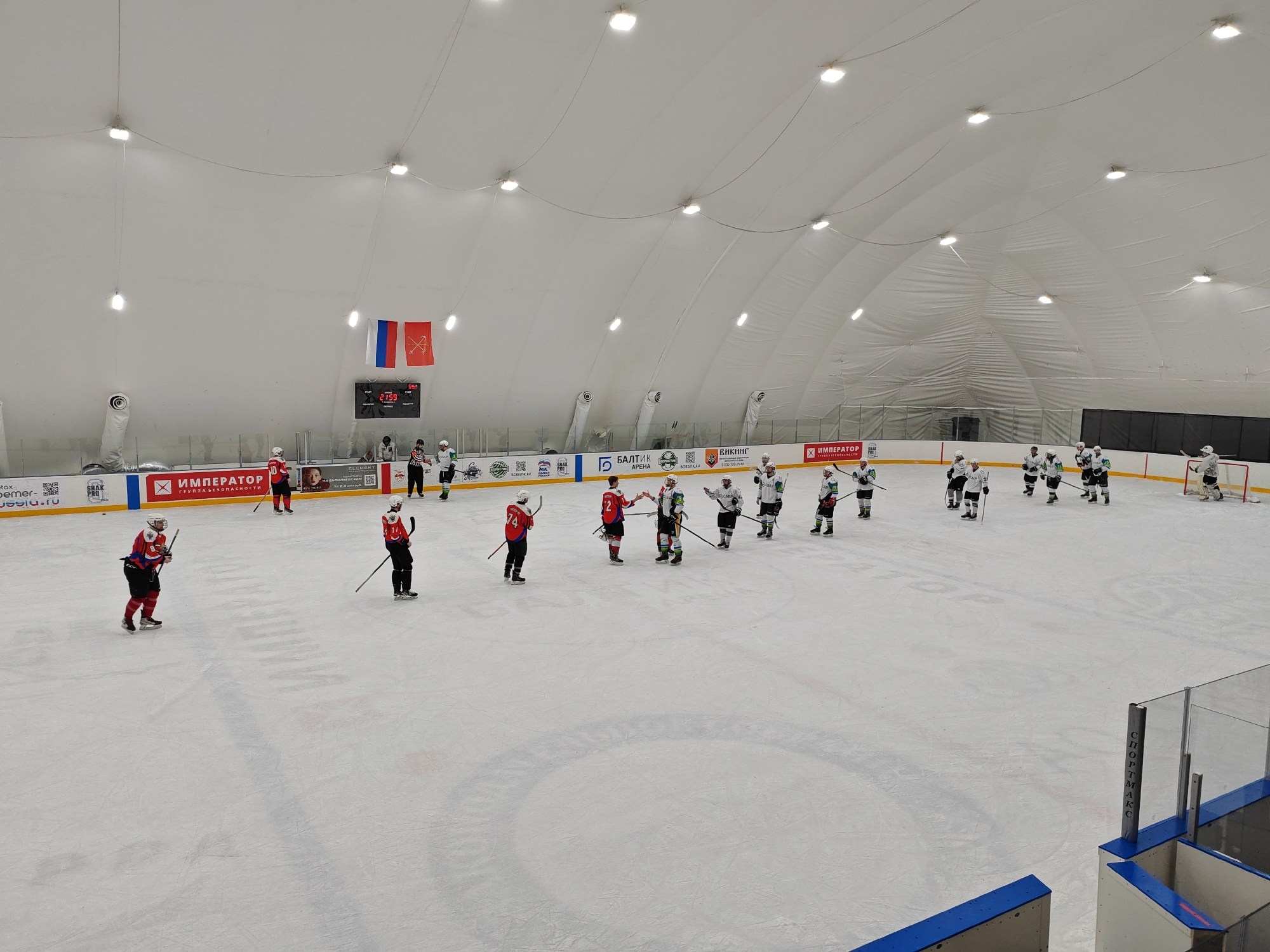 The image shows a hockey game in progress. The teams are wearing red and green uniforms. The players are standing on the ice and some are giving each other high fives. The environment is an indoor ice rink. There are several advertisements visible on the wall around the rink, as well as the Russian flag.