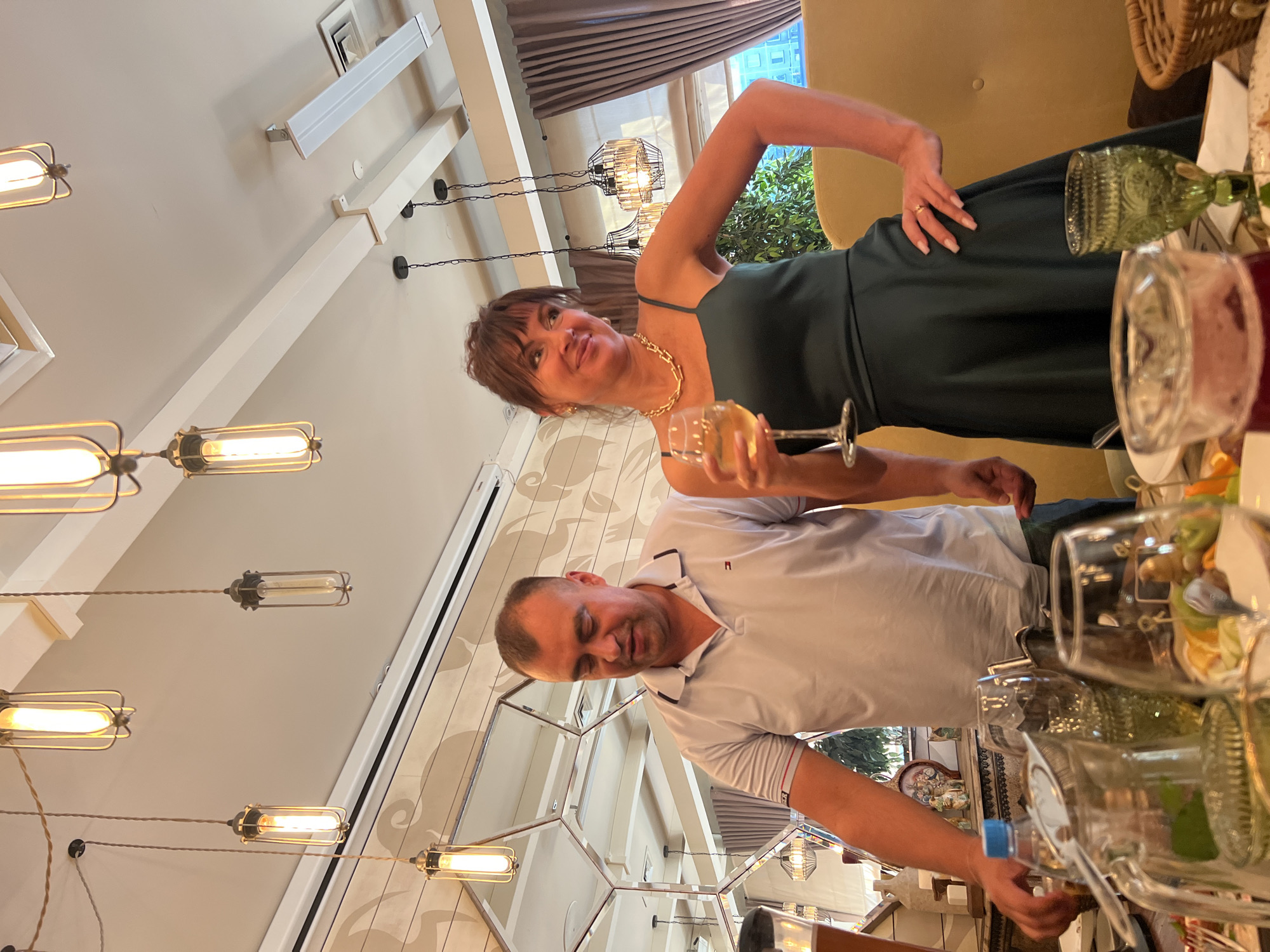 The image shows a man and a woman in a restaurant. The man is wearing a white polo shirt, and the woman is wearing a dark green dress and a gold necklace. The woman is holding a glass of white wine and smiling. The man is looking down, and his hands are on the table. There are several glasses and pitchers on the table, suggesting a meal is in progress. The restaurant appears to have a modern decor with hanging light fixtures. The scene is likely set in the city of Novosibirsk, Russia.