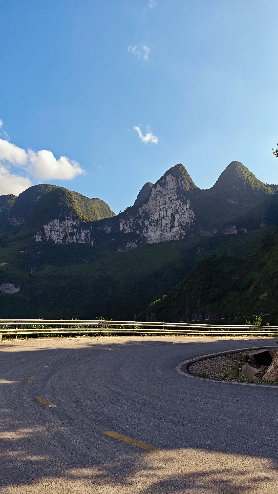 The image shows a curved section of asphalt road in Đồng Văn Commune, Vietnam. The road has yellow lane markings and a white guardrail. Mountains with visible rock formations and green vegetation are in the background. The sky is blue with scattered clouds. Shadows are visible on the road surface.